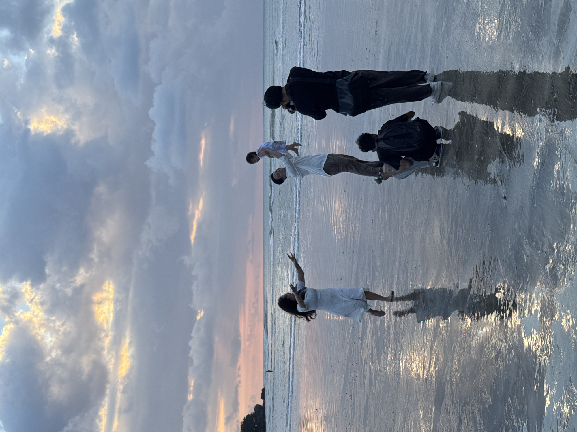 The image captures a serene scene on a beach in Kuta Selatan, Indonesia, at what appears to be golden hour, either sunrise or sunset. The sky is adorned with dramatic clouds, illuminated by warm hues of orange, pink, and yellow light breaking through. The wet sand on the shoreline beautifully reflects the sky and the subjects, creating shimmering patterns.
In the foreground, a woman in a white dress stands barefoot with her arms raised and slightly bent, her dark hair flowing. She appears to be looking towards the sky or the horizon. Further to the right, a man in a white shirt and light pants holds a baby, dressed in a light patterned outfit, playfully lifted in his arms. Both the man and baby seem to be engaging with each other or looking upwards.
Two other individuals are present, actively involved in photography. One man, dressed in a black jacket and dark pants, stands behind the man and baby, holding a camera to his eye, capturing the moment. Another person is partially visible, crouched down with a dark backpack, also appearing to be taking photos or videos from a lower angle.
The overall scene depicts a joyful group, likely a family or friends, enjoying a picturesque moment on the beach, possibly during a photoshoot. The atmosphere is peaceful and celebratory, highlighted by the stunning natural light and reflective wet sand. There is no visible text in the image.