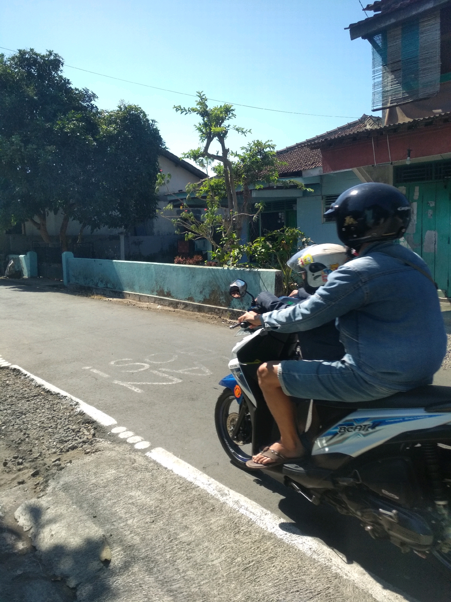The image shows a side view of two people on a scooter riding on a paved road in Desa Pamijen, Kecamatan Sokaraja, Indonesia. The person in the front is driving and wearing a denim jacket and shorts, with a black helmet. The person in the back is wearing a white helmet and a dark shirt. They are riding past a low, light blue wall with some greenery behind it. The road has white markings and a gravel shoulder. In the background, there are houses with tiled roofs and trees. The sky is clear and blue. The lighting suggests it is daytime.