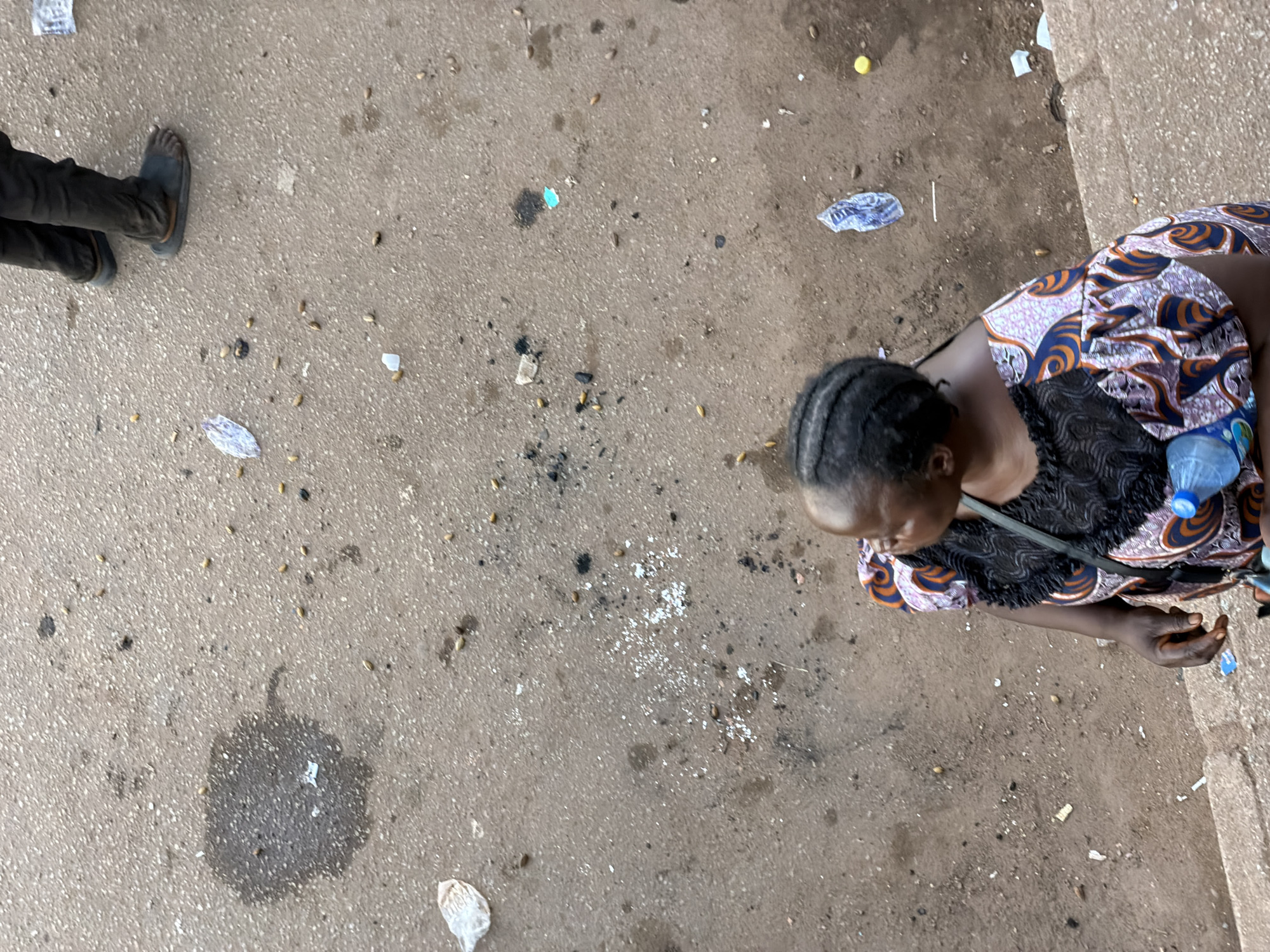 A high-angle, medium shot captures a woman standing on a sidewalk with her back to the camera, looking to her right. She has braided dark hair and is wearing a colorful, patterned dress with black trim. A dark strap is visible across her shoulder. She is holding a plastic water bottle with a blue cap. The sidewalk is made of concrete and is littered with debris including small pieces of trash, seeds, and what appears to be a dark stain from a spill. In the background, legs of another person wearing dark pants and sandals are visible. The scene is set on a sidewalk in Bukuru, Nigeria.