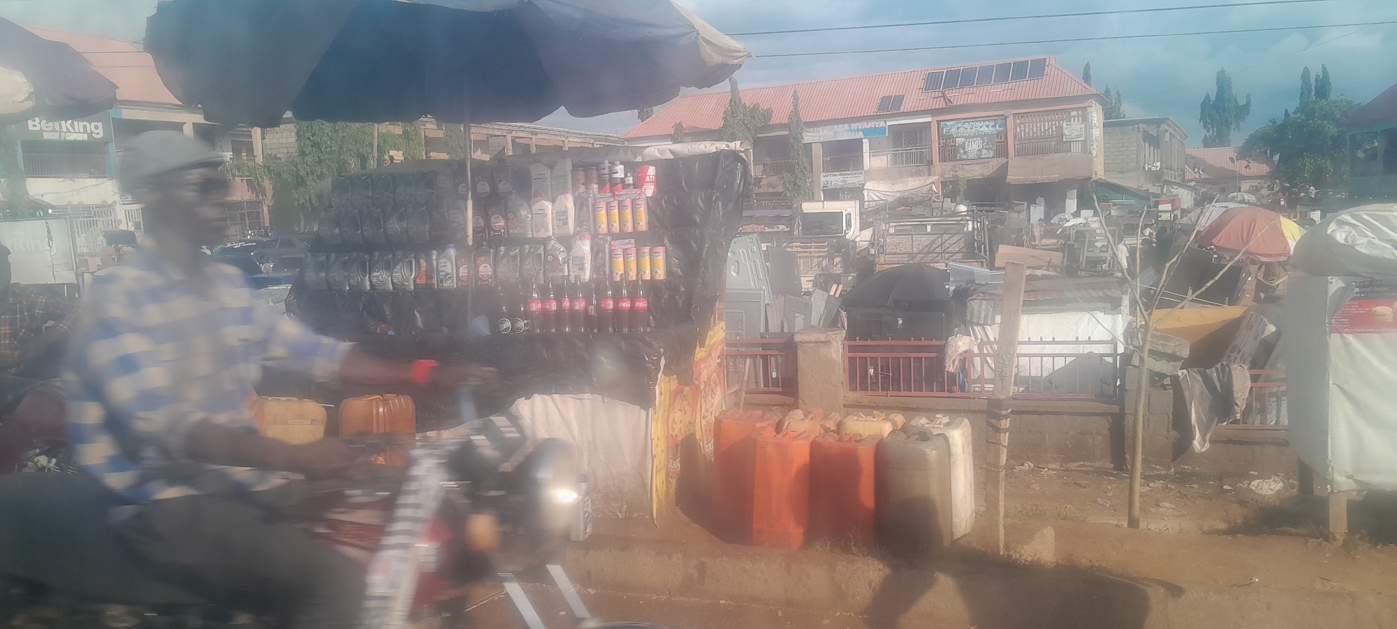 The image depicts a street scene in Nyanya, Nigeria, focusing on a roadside stall. A person wearing a checkered shirt and a cap is partially visible on a motorcycle in the foreground, appearing to be a vendor or customer. Behind this individual is a stall displaying a variety of items, including numerous bottles of what appears to be engine oil or similar automotive fluids, arranged on shelves. Below these bottles, several Coca-Cola bottles are visible. To the right of the stall, a collection of large plastic containers, some orange and some lighter in color, are stacked. The background reveals a densely packed urban environment with buildings, some featuring solar panels on their roofs. Trees and various structures, including what looks like other stalls or shops, are also present. The overall impression is of a bustling marketplace or commercial area.