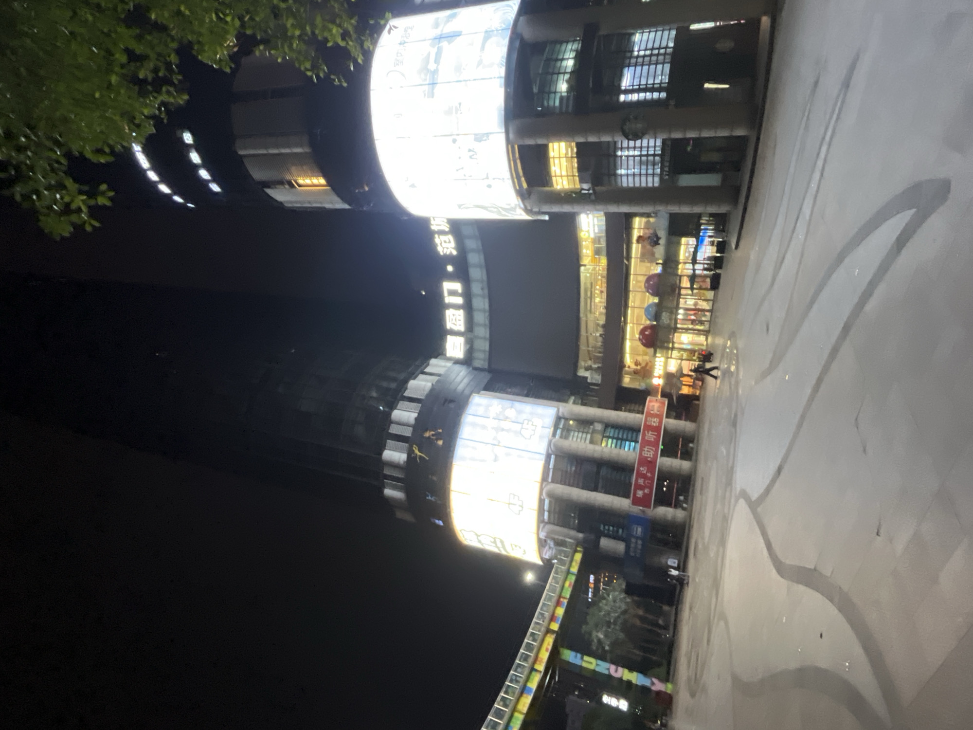 The image shows a nighttime view of a commercial district in Jiulongpo District, China. A large, modern building with illuminated signage and multiple floors dominates the scene. The building features a curved facade with large circular screens displaying advertisements and information. Adjacent to it is a taller, darker skyscraper. In the foreground, a wide, paved plaza is visible, with patterned lighting on the ground. Several people are present, walking across the plaza and in front of the building. A colorful elevated walkway or bridge is visible on the left side of the image, with the word "FUN CITY" illuminated on it. Green foliage from trees overhangs the top right corner of the image. The overall impression is a vibrant urban environment at night.