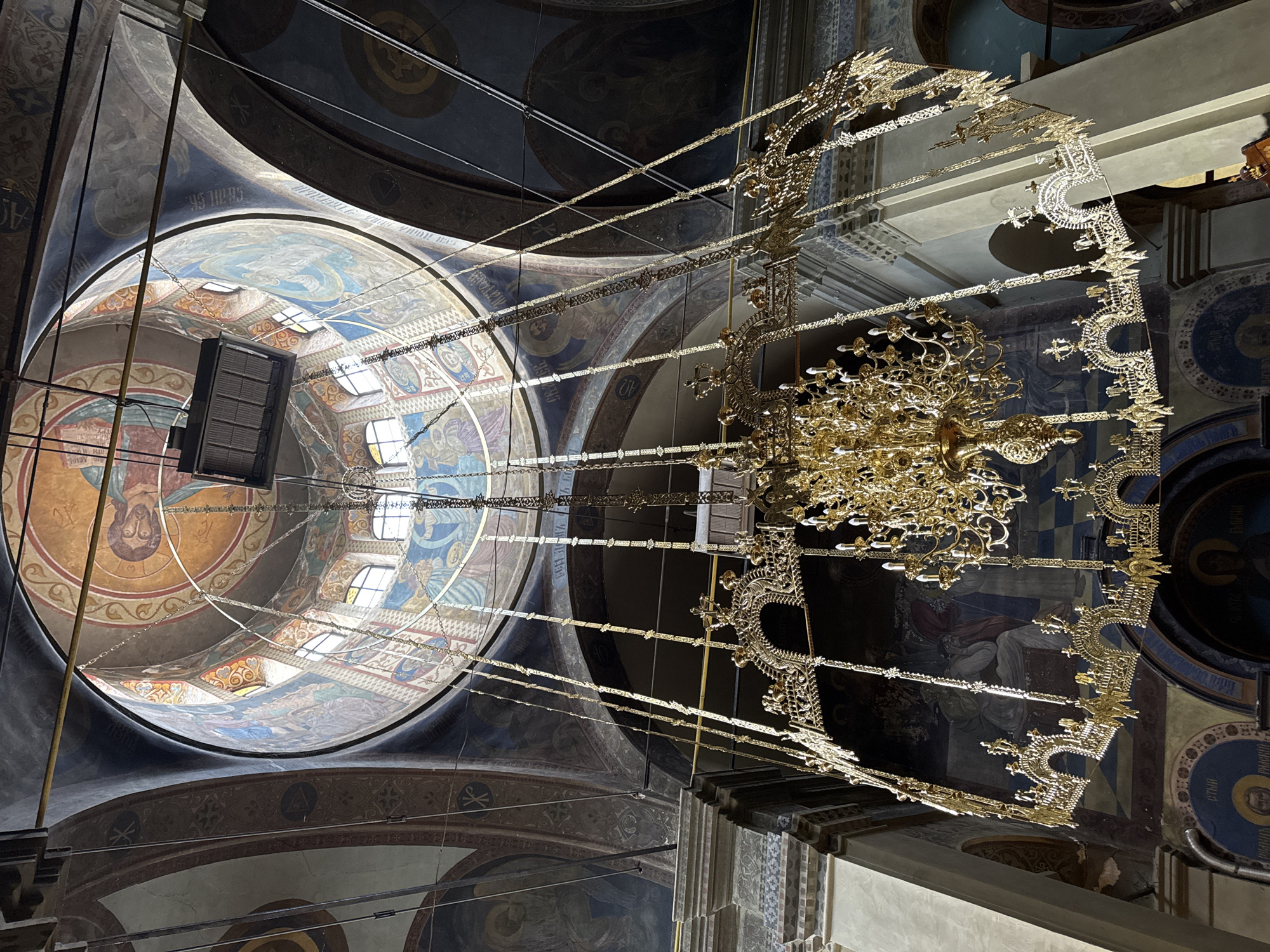 The image shows the interior of a church with a large, ornate golden chandelier hanging from the ceiling. The ceiling is adorned with colorful frescoes depicting religious figures and scenes. Light streams in from windows in the dome, illuminating the chandelier and the artwork. The walls also feature painted murals and architectural details like columns and arches. The perspective is looking upwards from a low angle, emphasizing the grandeur of the space and the intricate details of the chandelier and ceiling decorations. The church appears to be a historical and possibly Orthodox Christian site. While specific location details like the city of Yambol, Bulgaria are not visible in the image itself, this information has been provided as context. There are no people visible in the image.