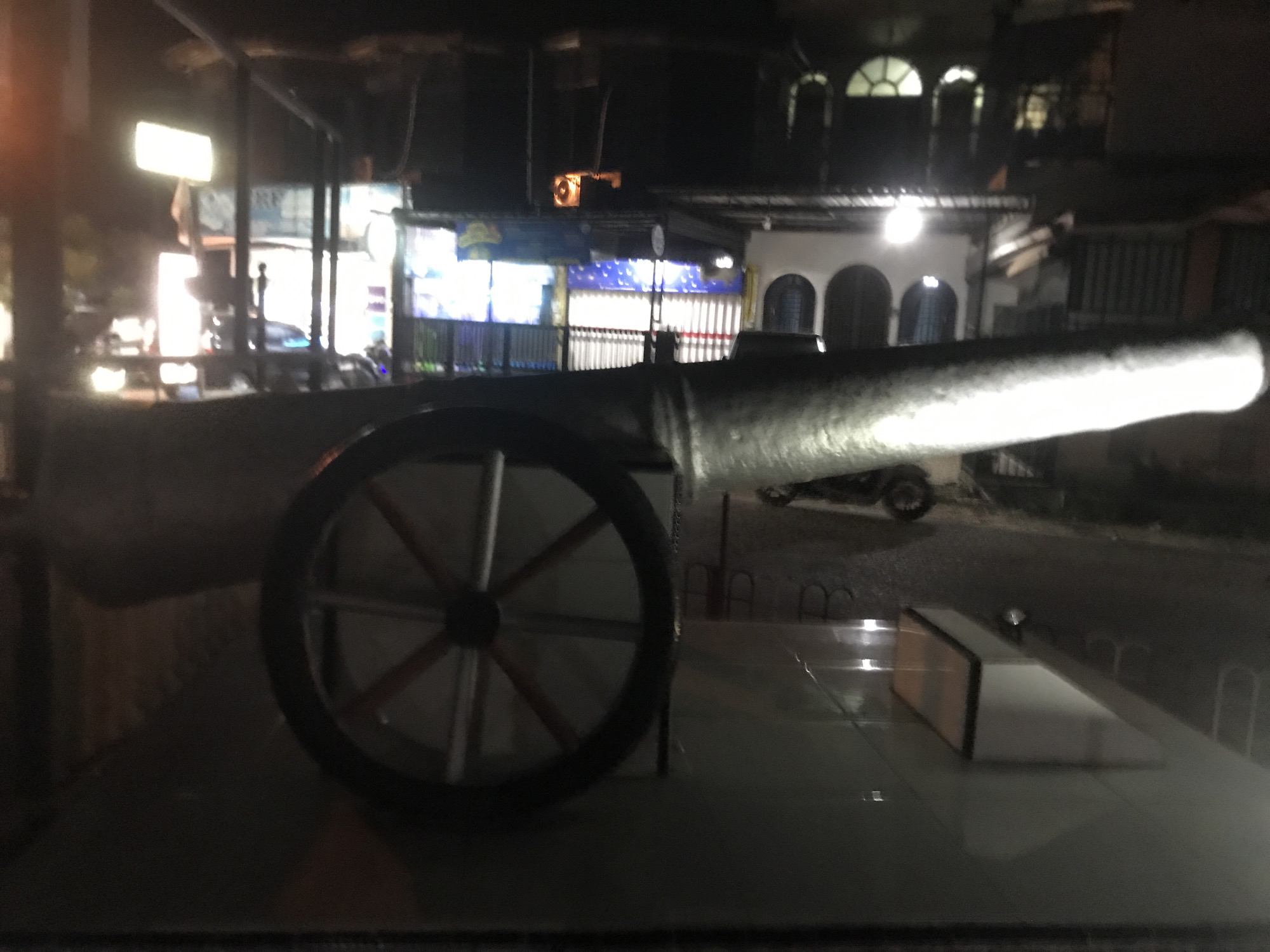 The image depicts an old cannon exhibited in Lhokseumawe, Indonesia, likely as a historical artifact or monument. The cannon is mounted on a wheeled carriage with wooden wheels and is positioned on a platform, possibly for display purposes. The background features dimly lit buildings and streetlights, indicating that the photograph was taken at night. The scene appears to be in a public area within the city.
