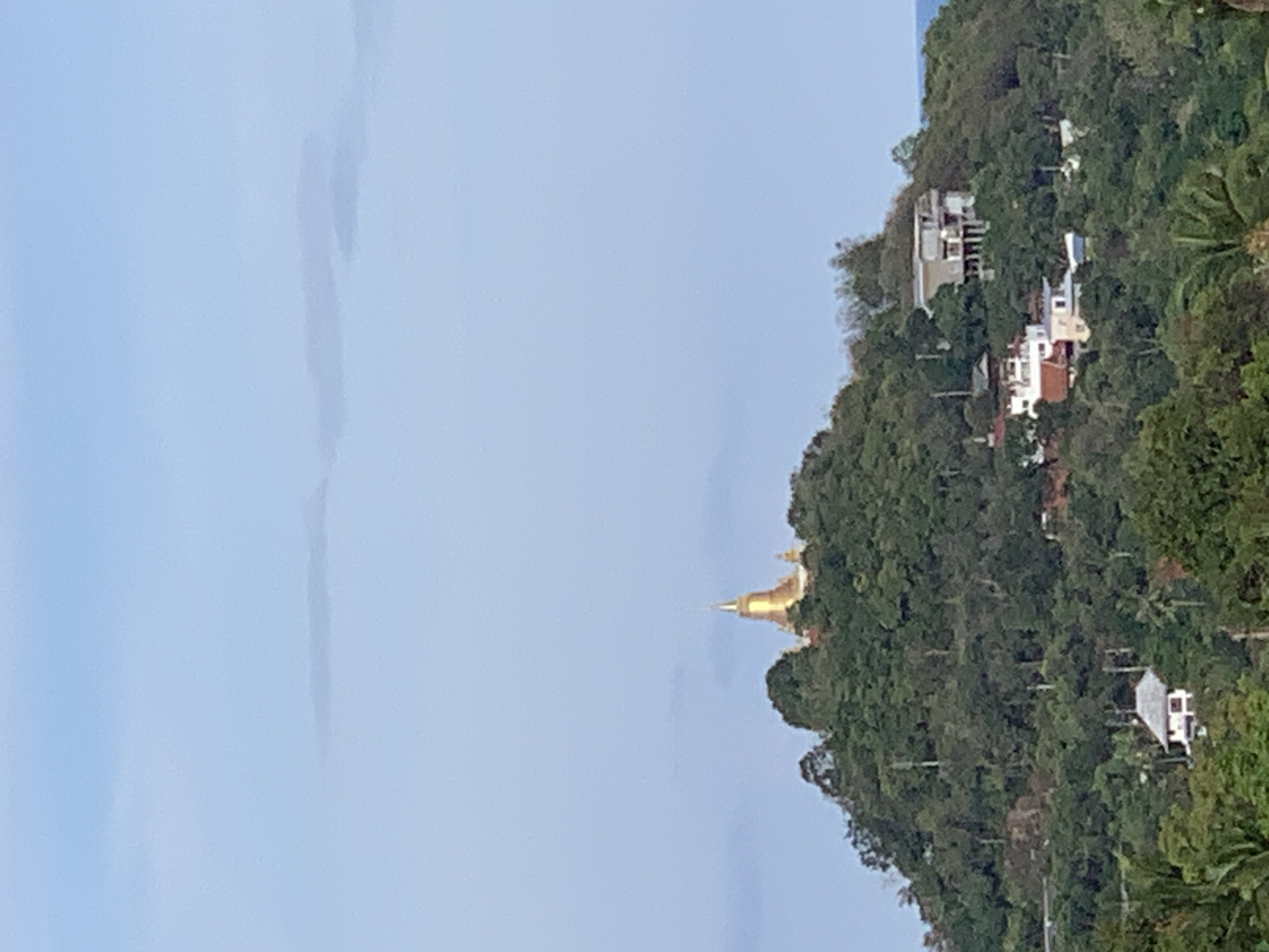 The image captures a view from an elevated perspective in the city of บ้านบางรักษ์, Thailand. A large section of the frame is filled with a pale blue sky that shows evidence of cloud cover. Situated on the right is a lush, green hillside densely populated with trees and vegetation. Nestled within the trees are several buildings, including residential homes and a gleaming, golden Buddhist temple, which is the focal point. The buildings are varied in their architecture and design. The hillside slopes upwards, indicating it is part of a raised area.