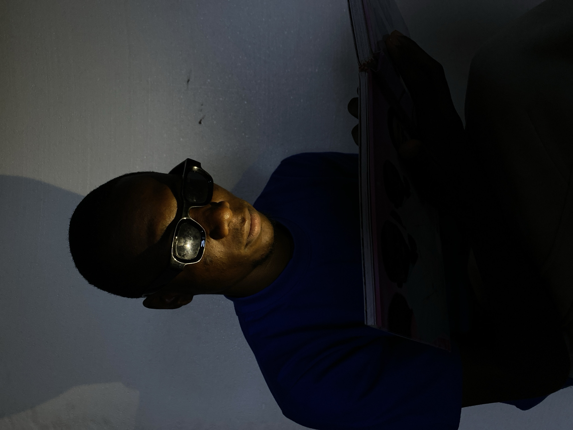 In a dimly lit interior, a young Black male wearing black-rimmed glasses and a royal blue t-shirt is engrossed in reading a book. The book is held open, its pages illuminated by a light source that casts shadows on the man's face and the surrounding wall. The wall, textured and appearing to be a light grey or white, shows subtle shadows and possibly some imperfections. The man's gaze is directed downwards, focused on the content of the book. The overall scene suggests a quiet moment of study or reading in a low-light environment.