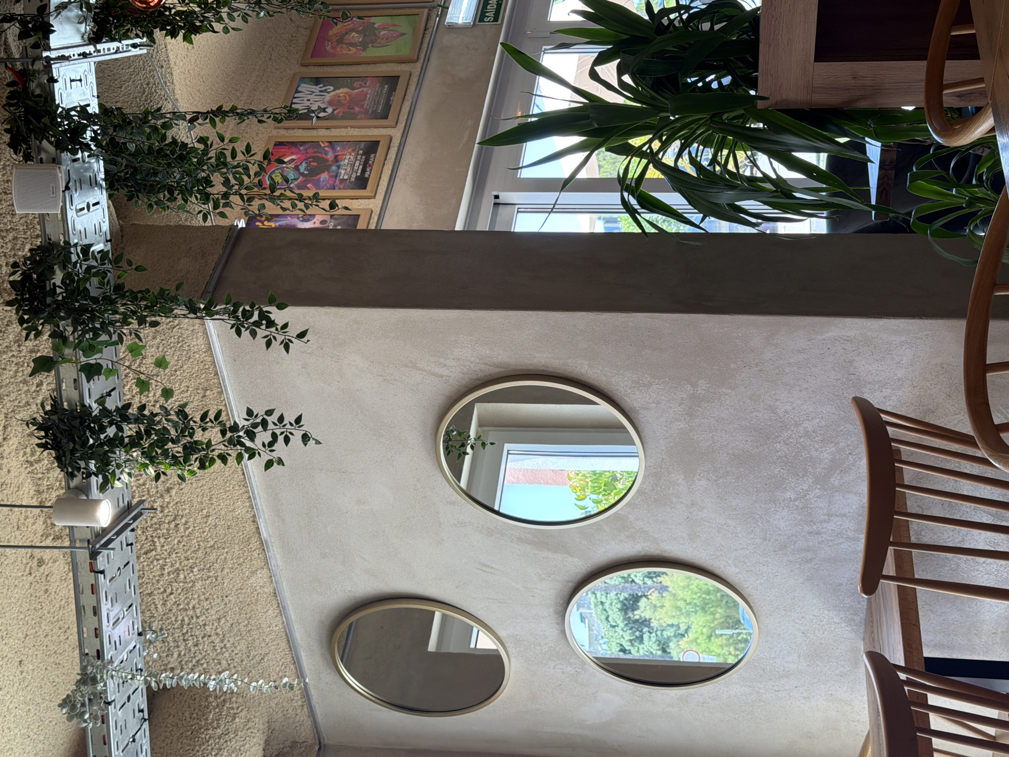 The image shows an interior scene with a variety of elements. In the foreground, we see three oval mirrors mounted on a textured concrete wall. These mirrors reflect a window, greenery, and parts of the ceiling. To the right, a portion of a wooden chair with a slatted back is visible.
In the background, hanging plants with lush green leaves cascade down from above, attached to a metal shelving unit. These plants add a touch of nature to the indoor environment. Also visible in the background are framed posters or artworks displayed on a wall. One poster appears to have a title that might be "MAXIM." A white speaker or light fixture is mounted on the metal shelving.
The scene suggests a cafe, restaurant, or a similar establishment, possibly with a bohemian or eclectic design aesthetic. The presence of framed posters and hanging plants hints at a creative or artistic atmosphere. The reflections in the mirrors indicate natural light coming from a window, suggesting it's daytime. The overall impression is one of a well-decorated and inviting space. No people are visible in the image. The location context of Cascais, Portugal, provides a geographical anchor for this scene.
