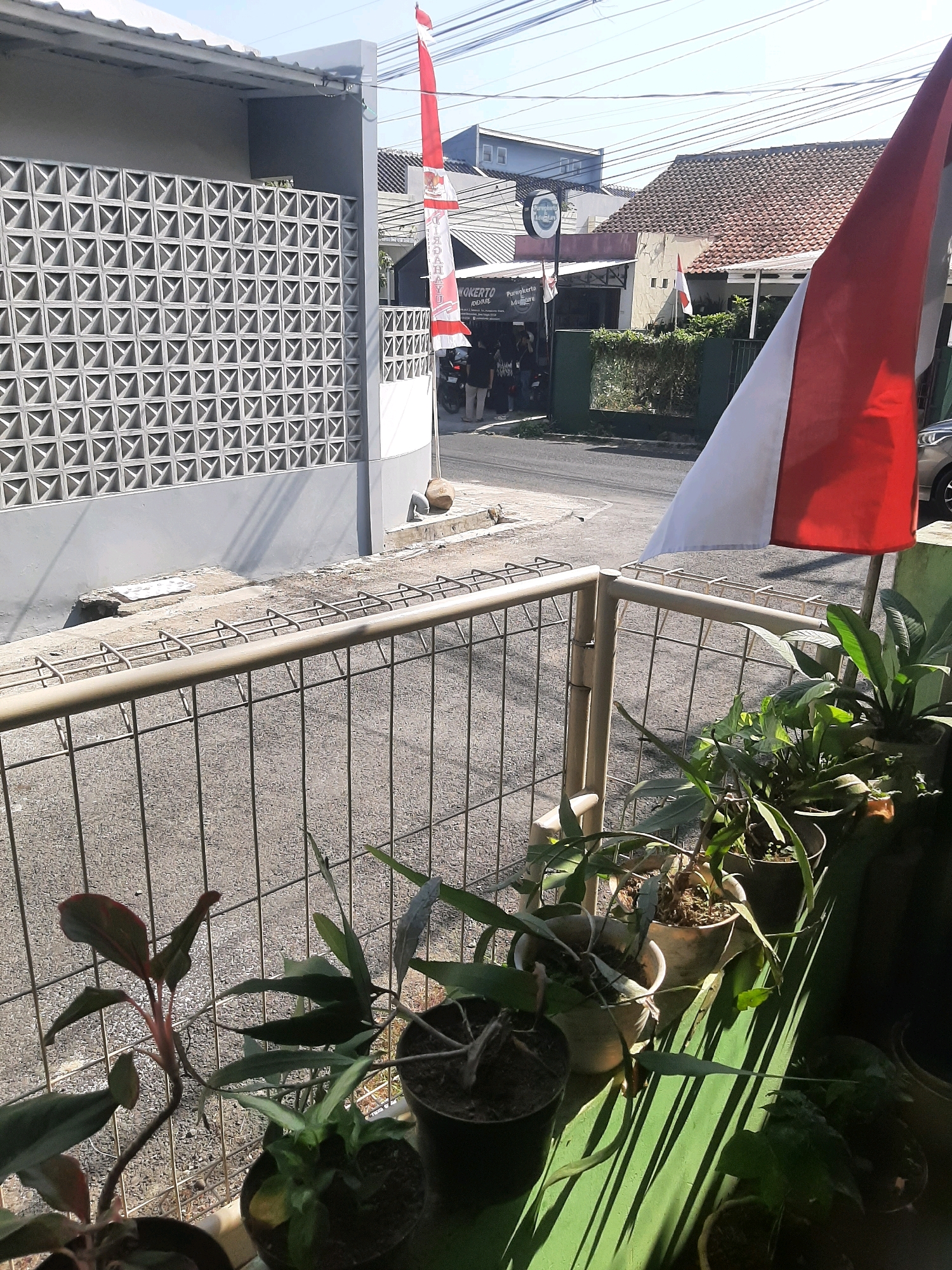 A narrow shot taken from a balcony shows a street scene in Sumampir, Indonesia. In the foreground, a metal fence with a light brown rail encloses potted plants, some of which are in shadow. To the right, an Indonesian flag, red and white, hangs prominently. Beyond the fence, a street is visible, with a gray wall with a decorative square pattern to the left. A building with a red-tiled roof stands across the street, with a circular sign that reads "Purwokerto Adventure". Several people are gathered in front of this building. A car is parked on the right side of the street. The sky is clear and blue.