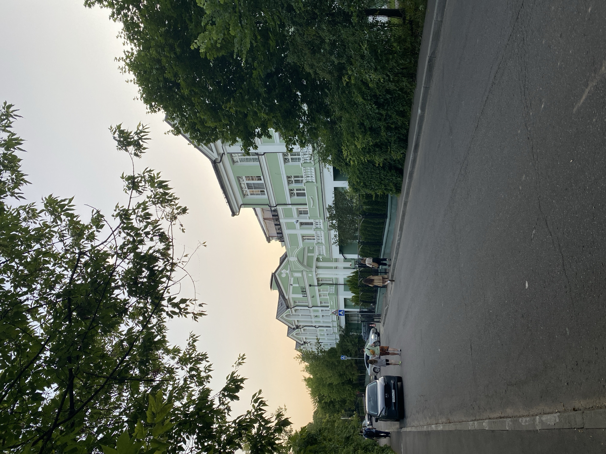 The image depicts a street scene in Khimki, Russia, likely during the late afternoon or early evening, given the soft, warm lighting and the gentle gradient of the sky from pale yellow to white. The focus is on a row of light green, multi-story buildings with white trim and numerous windows. The architecture suggests a residential or possibly a mixed-use area, with a formal, classic style. Lush green trees line the street, providing a canopy over parts of the sidewalk and obscuring some of the buildings.
A paved road runs diagonally across the frame, with a curb separating it from a green, tree-filled area. Several people are visible walking along the sidewalk next to the road and near the buildings. Some are casually strolling, while others appear to be conversing. A few cars are parked along the street and one is in motion. One car has a visible license plate that reads "925XA 777". There are also blue traffic signs visible on poles. The overall atmosphere seems peaceful and everyday, capturing a moment of urban life under a slightly overcast or hazy sky.