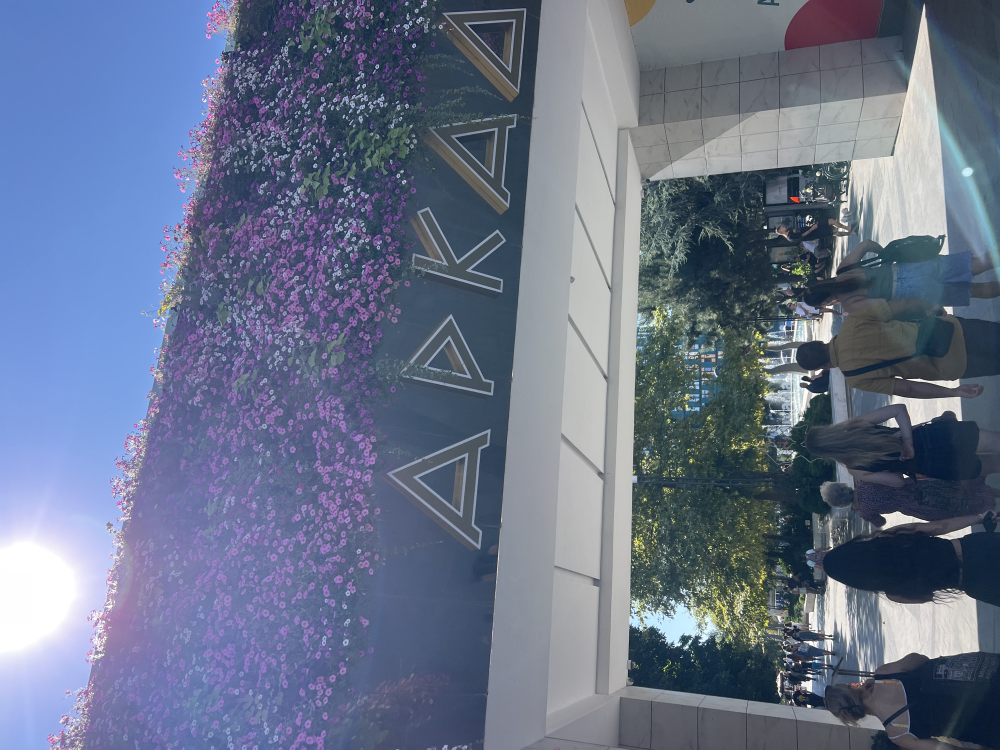 The image shows an outdoor scene in Odesa, Ukraine, under a bright sun. In the foreground, a group of people are walking away from the camera, framed by a modern white archway. Above the archway, a dark wall is covered with a dense cascade of pink and white flowers. Large, stylized golden letters with white outlines spell out "АРКАДІЯ" on the wall, indicating the location is Arcadia, a popular resort area in Odesa. Beyond the archway, a sunlit park is visible with trees, people strolling, and a fountain in the background. The overall impression is of a pleasant, lively day in a well-maintained public space.