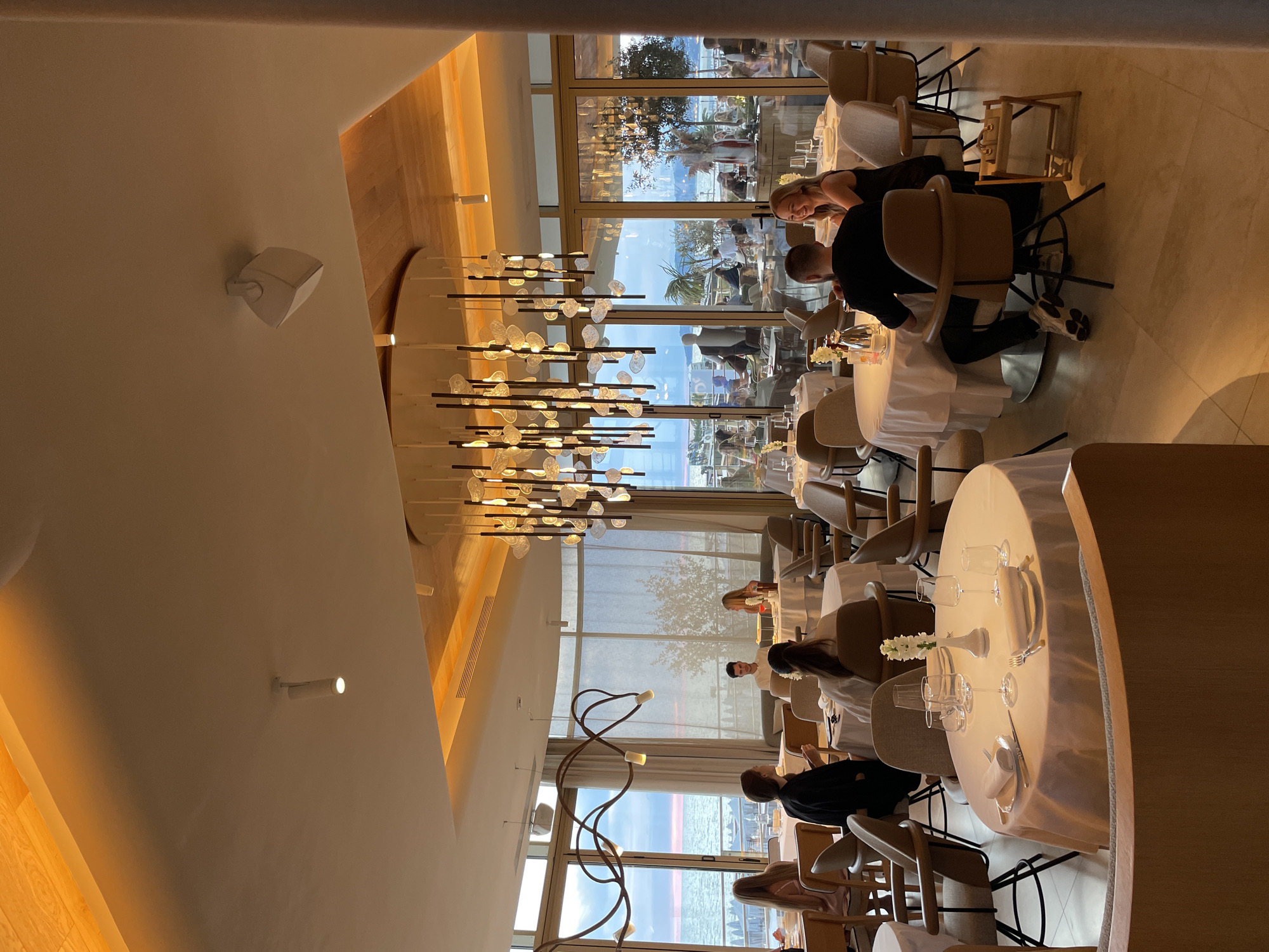 The image depicts the interior of a high-end restaurant with a view of the sea. The restaurant is illuminated by warm, ambient lighting from a large, multi-tiered chandelier and recessed ceiling lights. Several tables, covered with white tablecloths and set with fine china, are arranged throughout the space. Some tables are occupied by diners, including a couple in the foreground engaged in conversation. In the background, through large windows, a seascape with a sunset sky and a shoreline with beach umbrellas can be seen. The restaurant's design appears modern and elegant, with comfortable seating and decorative elements. It is not possible to determine the specific location within the city of Sochi, Russia, from the image alone.