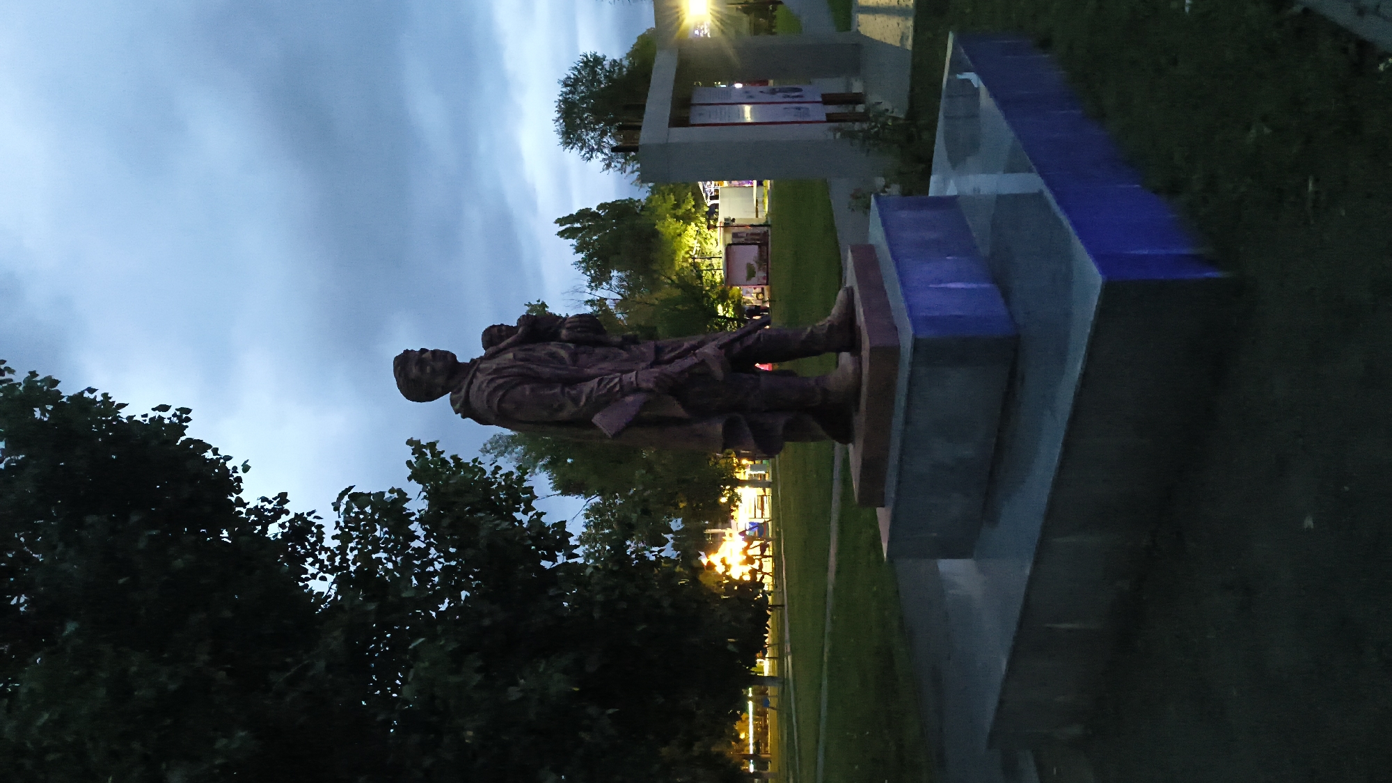 This is a full-shot, eye-level, vertical, medium-light, dusk photograph of a bronze statue in a park. The statue depicts a man wearing a long coat, carrying a child on his left shoulder. He is facing right in profile and holding a rifle in his right hand. The statue is mounted on a multi-tiered, gray, stone pedestal, which is illuminated from below with blue light. The statue is located in a park with green grass, trees, and a cloudy sky. In the background, there are lights from buildings and what appears to be a playground. The overall mood is somber due to the dusk lighting and the subject matter of the statue, which may represent a soldier or a figure from a historical event. This statue is located in the city of Mikhaylovka, Russia.