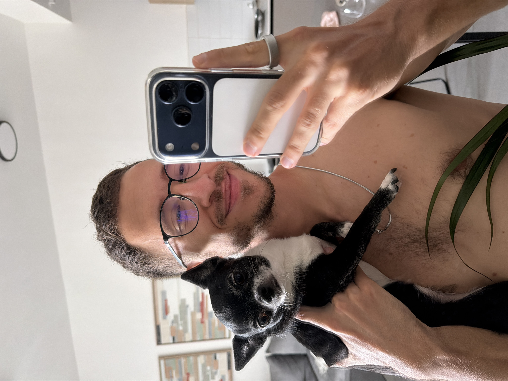 A man is taking a selfie with a black and white chihuahua dog. The man is wearing glasses and has a silver ring on his finger. He is shirtless. The location appears to be inside a building. It is not possible to determine if the location is in Tel-Aviv, Israel without further information.