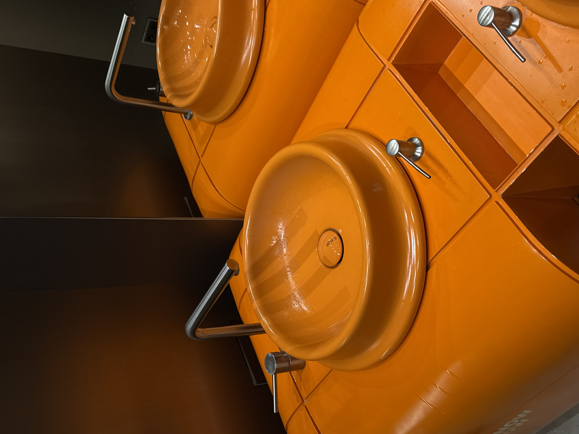 This image shows an orange washbasin with two circular sinks and integrated shelves. The fixtures are made of metal and have clean lines. It is a well-lit indoor setting, possibly a public restroom. The location is Yerevan, Armenia.