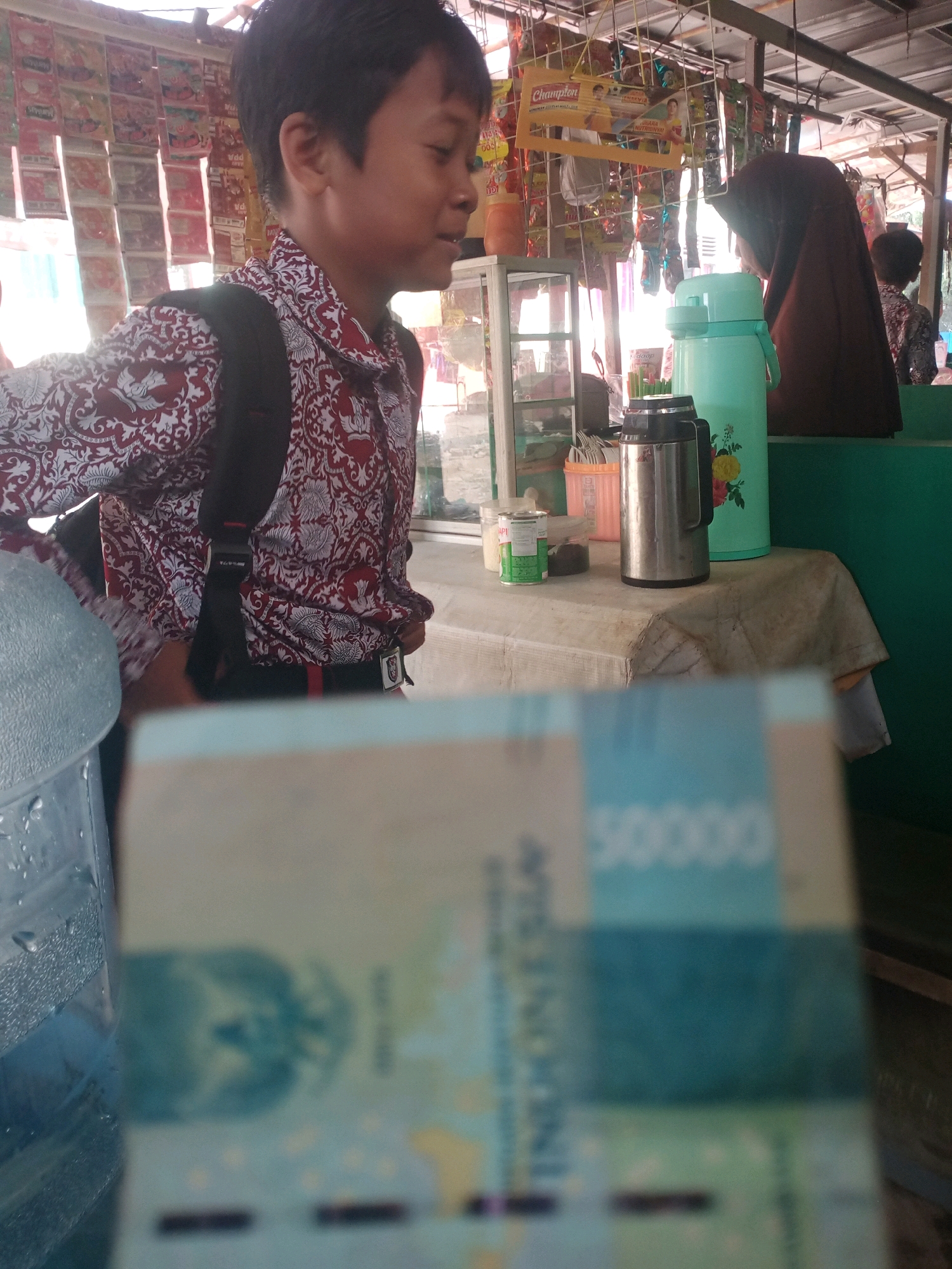 The image depicts an outdoor stall, likely in Bandar Lampung, Indonesia. In the foreground, a partially blurred banknote is visible, displaying the denomination "50000". To the left, a large blue water dispenser is present. The middle ground features a young boy wearing a red and white patterned shirt and a black backpack, looking to his right. Behind him, a woman wearing a brown hijab is seen partially from the side, looking downwards. A teal-colored thermos is on a table covered with a cloth, along with other items including a metal thermos and containers. The background shows a rack filled with various packaged goods, suggesting it is a retail setting. The environment appears to be a street-side stall with a simple roof structure.