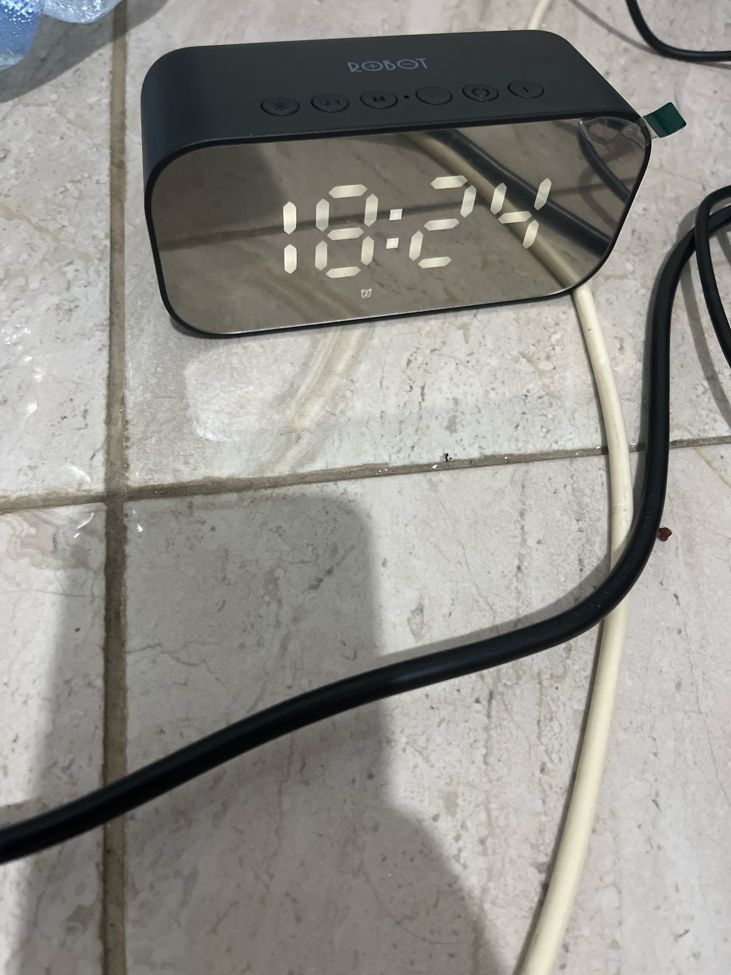 The image shows a black digital alarm clock with a mirrored LED display showing the time as 18:24. The clock has the brand name "ROBOT" printed on the top. It is placed on a light-colored tiled surface. Two electrical cords, one black and one white, are visible near the clock, suggesting it is plugged in. The overall scene appears to be an indoor setting, possibly a room or floor, with the tiles showing some signs of wear or dirt. There are no people visible in the image. The location is not specified, but the query mentions Matesih, Indonesia, which is a subdistrict in Central Java.