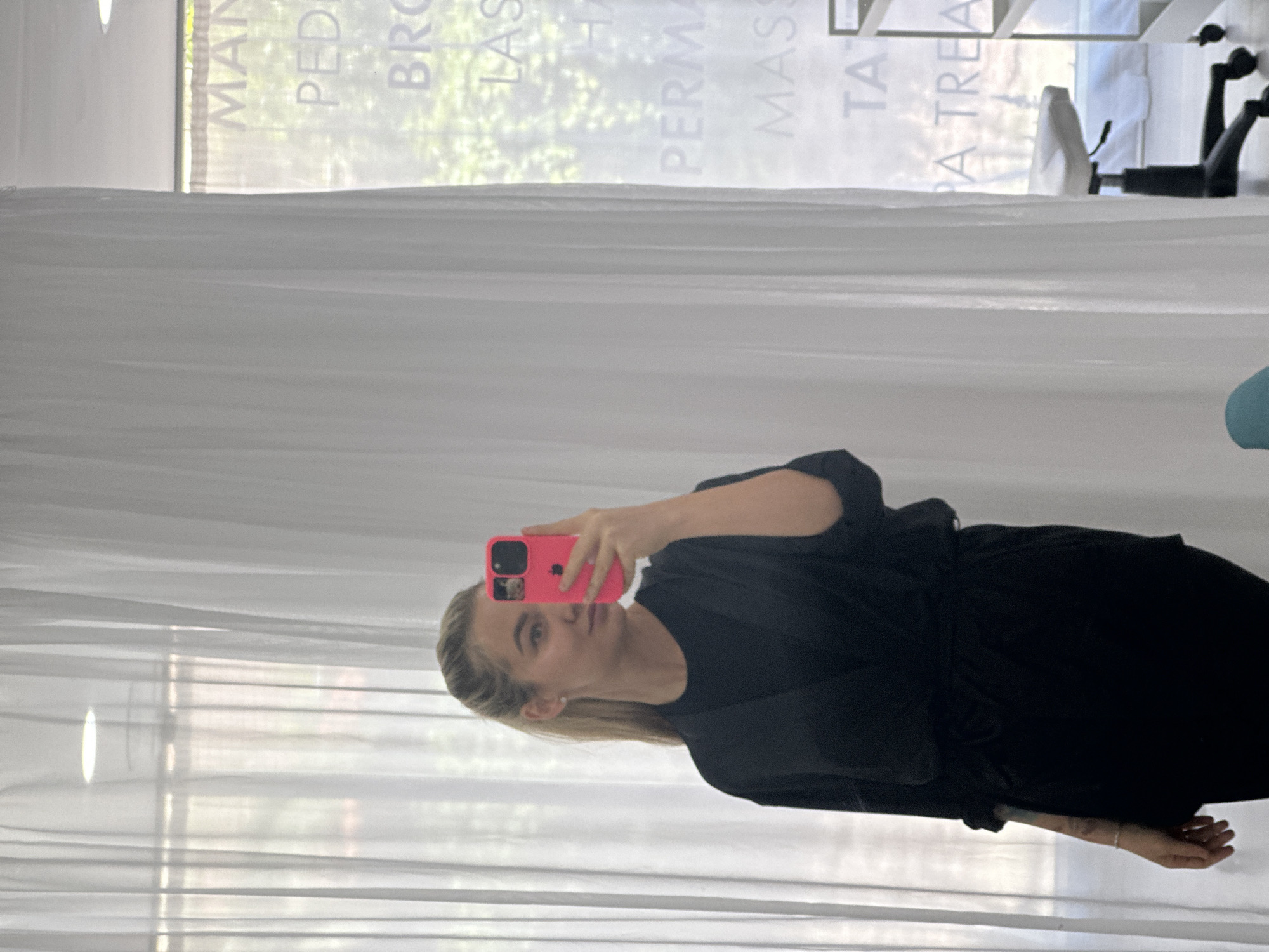 A woman with blonde hair, wearing a black outfit, is captured in a selfie taken indoors, looking directly at the camera. She holds a bright pink smartphone, with a distinctive camera module, in her right hand to take the picture. The scene is set against a large window, covered by sheer white curtains that extend from floor to ceiling.
Through the window and reflected on its surface, various words associated with beauty and wellness services are visible, though reversed and slightly blurry. Decipherable words include "MANICURE" (partial), "PEDICURE", "BROWS", "LASHES", "PERMANENT MAKEUP", "MASSAGE", "TATTOOS", and "SPA TREATMENT". These texts suggest the location is a salon, spa, or similar establishment in Budva, Montenegro.
Outside the window, green foliage is discernible, indicating a view of trees or bushes and suggesting it is daytime. An office-style chair with wheels and a light-colored surface or table are also visible in the background on the right side. The woman has a neutral expression, and a subtle tattoo can be seen on her lower left arm. The overall lighting is soft, diffused by the white curtains.