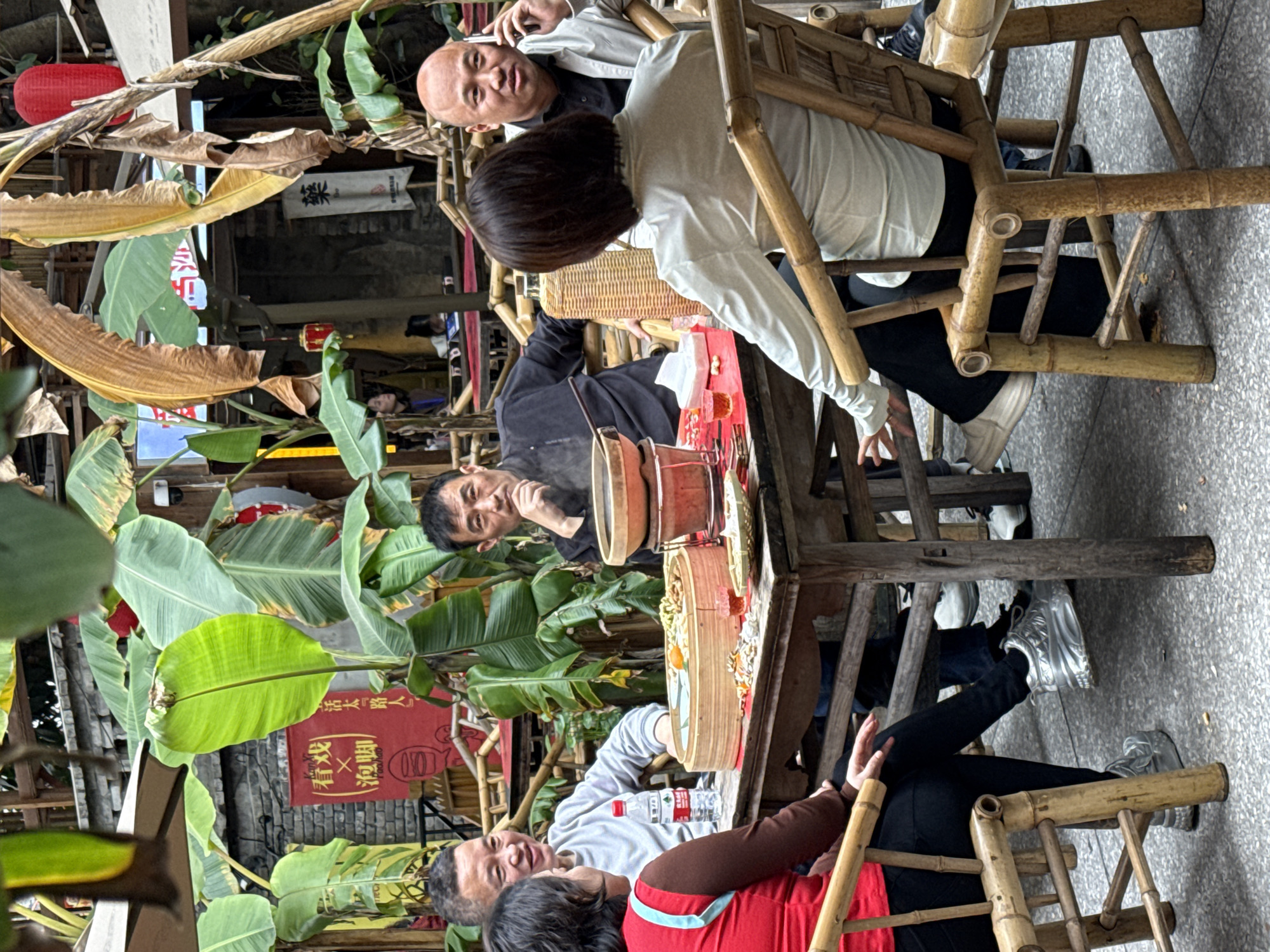Five individuals, four men and one woman, are seated around a square wooden table in an outdoor or semi-outdoor restaurant setting within Yuzhong District, China. The environment is characterized by abundant large green and dried banana leaves, indicating a tropical or subtropical ambiance. In the foreground, a woman in a red vest over a dark top and black pants, wearing silver platform shoes, is seated on a bamboo chair, partially facing the camera. To her right, a man in a light grey long-sleeved shirt is smiling and looking towards the woman, with a plastic water bottle on the table in front of him. Opposite them, a man in a dark long-sleeved shirt observes the scene with his hand to his chin. In front of him is a round clay or ceramic hotpot containing a steaming substance, positioned next to a large round bamboo steamer filled with various food items. To the right, another woman, with her back mostly to the camera, wears a light-colored long-sleeved top and dark pants, seated on a bamboo chair. Beside her, a bald man in a light jacket is actively engaged in a phone call, holding a mobile phone to his right ear. The table also holds small red drinking glasses, a tissue box, and a small woven bamboo plate. The ground beneath the table is a light-colored, speckled pavement. In the background, brick walls and wooden structures are visible, adorned with red lanterns and two banners: one red banner displays Chinese characters including "看戏 X 泡脚" (Watch Opera X Foot Bath), and a white banner shows the character "榮" (Róng).