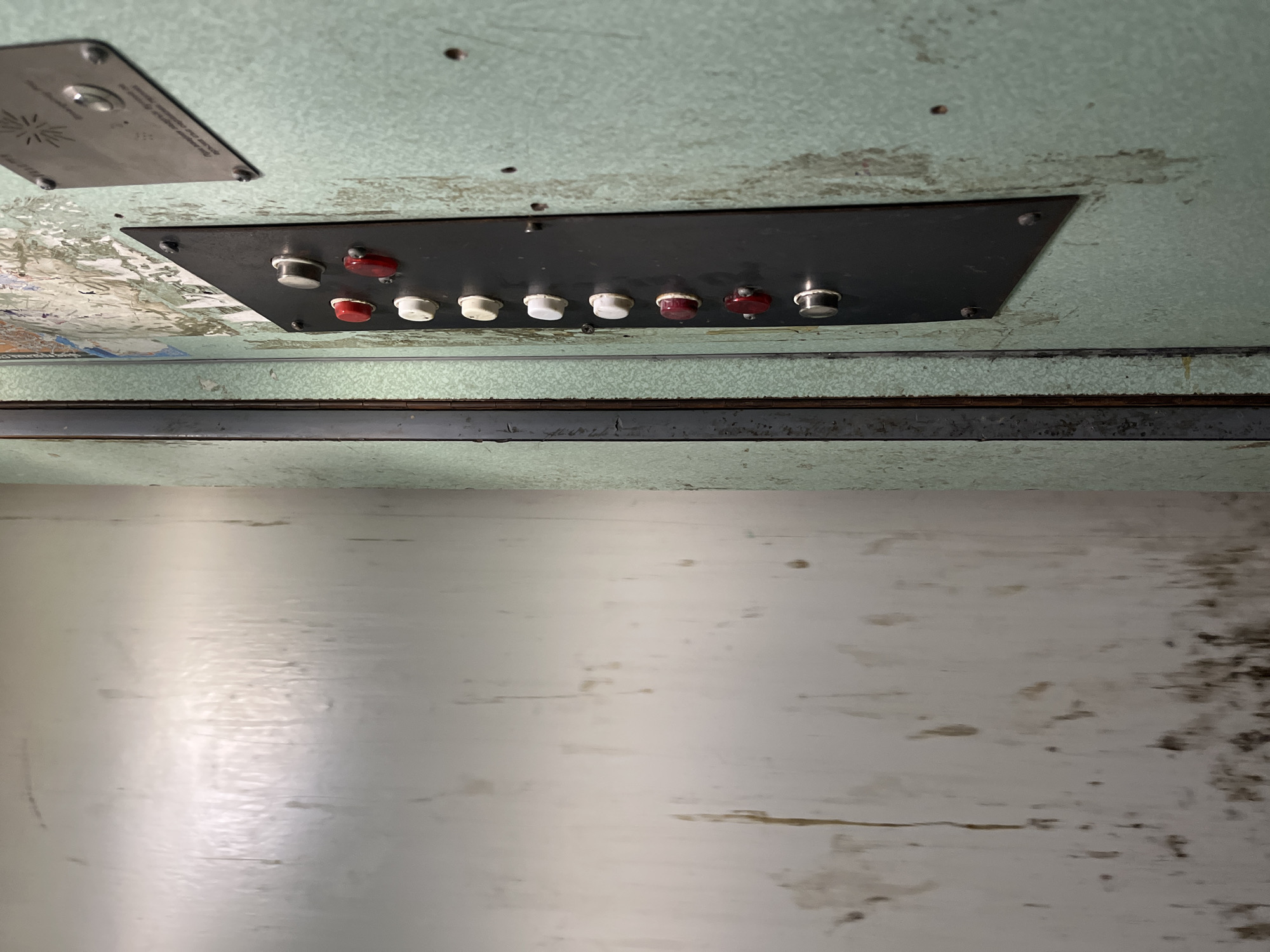 The image shows a close-up of the interior ceiling of a vehicle, likely a public transport vehicle such as a bus or tram. The ceiling has a light green patterned surface, which is weathered and shows signs of wear and tear. A black rectangular panel is mounted on the ceiling and contains a row of eight push buttons, alternating between white and red. A smaller, metallic plate with symbols is also affixed to the ceiling near the button panel. Below the ceiling is a grey metal bar, and below that is a large, light-colored, worn surface that appears to be a wall or panel. The overall impression is one of an aged and somewhat neglected interior space. The location in Sofia, Bulgaria, cannot be definitively determined from the image alone.