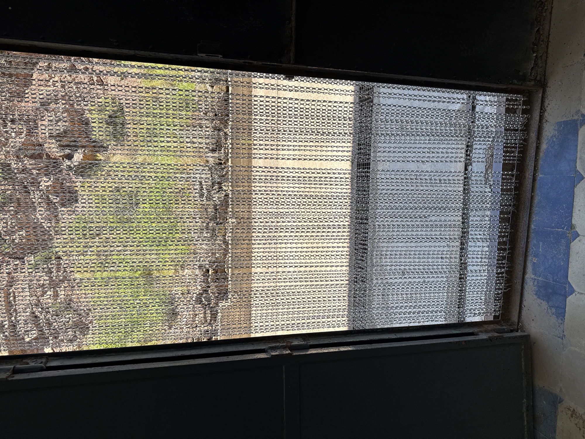 The image shows an open, dark metal doorway with a chain-link curtain hanging in the opening. The doorway leads to an outdoor area with rocky, uneven terrain and patches of green vegetation visible through the chain curtain. The ground inside the doorway is tiled with a pattern of blue and light-colored squares. The chain curtain is made of interlocking metal rings, creating a textured, semi-transparent barrier. The overall scene suggests an entrance to a natural or semi-abandoned area.