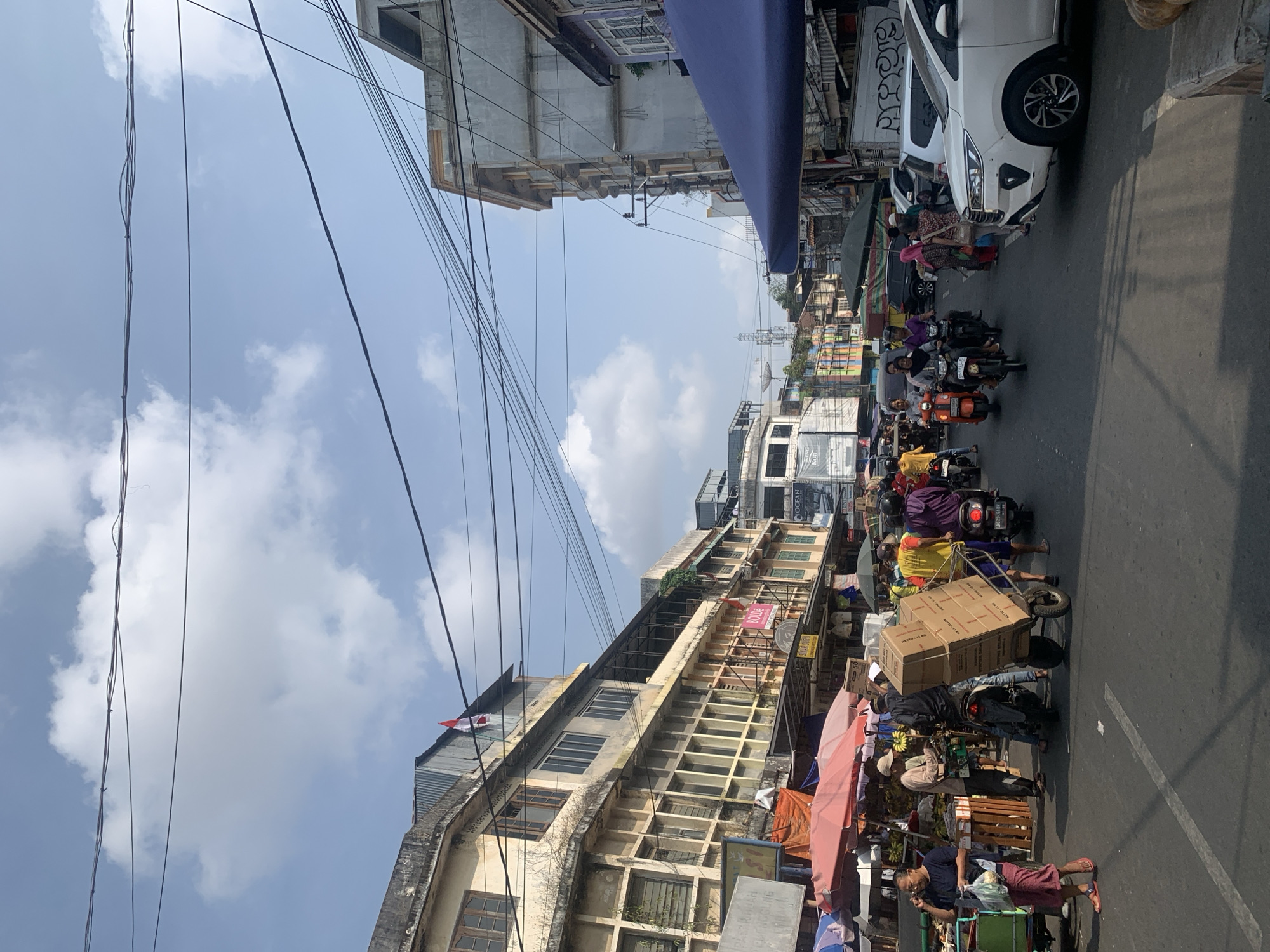 The image depicts a bustling street scene in Palembang, Indonesia, likely a marketplace area given the presence of vendors, goods, and various modes of transportation. The perspective is from ground level, looking down a moderately busy street.
In the foreground, a white SUV is parked on the right side of the road. To the left, a woman in a blue shirt and patterned skirt walks with a plastic bag in her left hand, speaking on her phone. She is standing next to a green cart laden with what appears to be produce. Behind her, a man in a light brown shirt and cap is interacting with another man. A motorcycle is parked next to them, and a cart loaded with several cardboard boxes is being pushed or pulled by a person. More people, some on motorcycles, are visible further down the street.
The street is lined with multi-story buildings on both sides, some of which appear older and somewhat weathered, with exposed pipes and signs in Indonesian. One building on the left has a red and white Indonesian flag displayed. Several stalls with colorful awnings and umbrellas are set up along the sidewalks, suggesting a market environment.
The sky is blue with scattered white clouds, and the overall lighting suggests it is daytime. Overhead, a complex network of electrical wires crisscrosses the sky, typical of urban environments in Indonesia. The traffic consists of motorcycles, a few cars, and possibly some pedestrians pushing carts. The atmosphere appears active and vibrant.