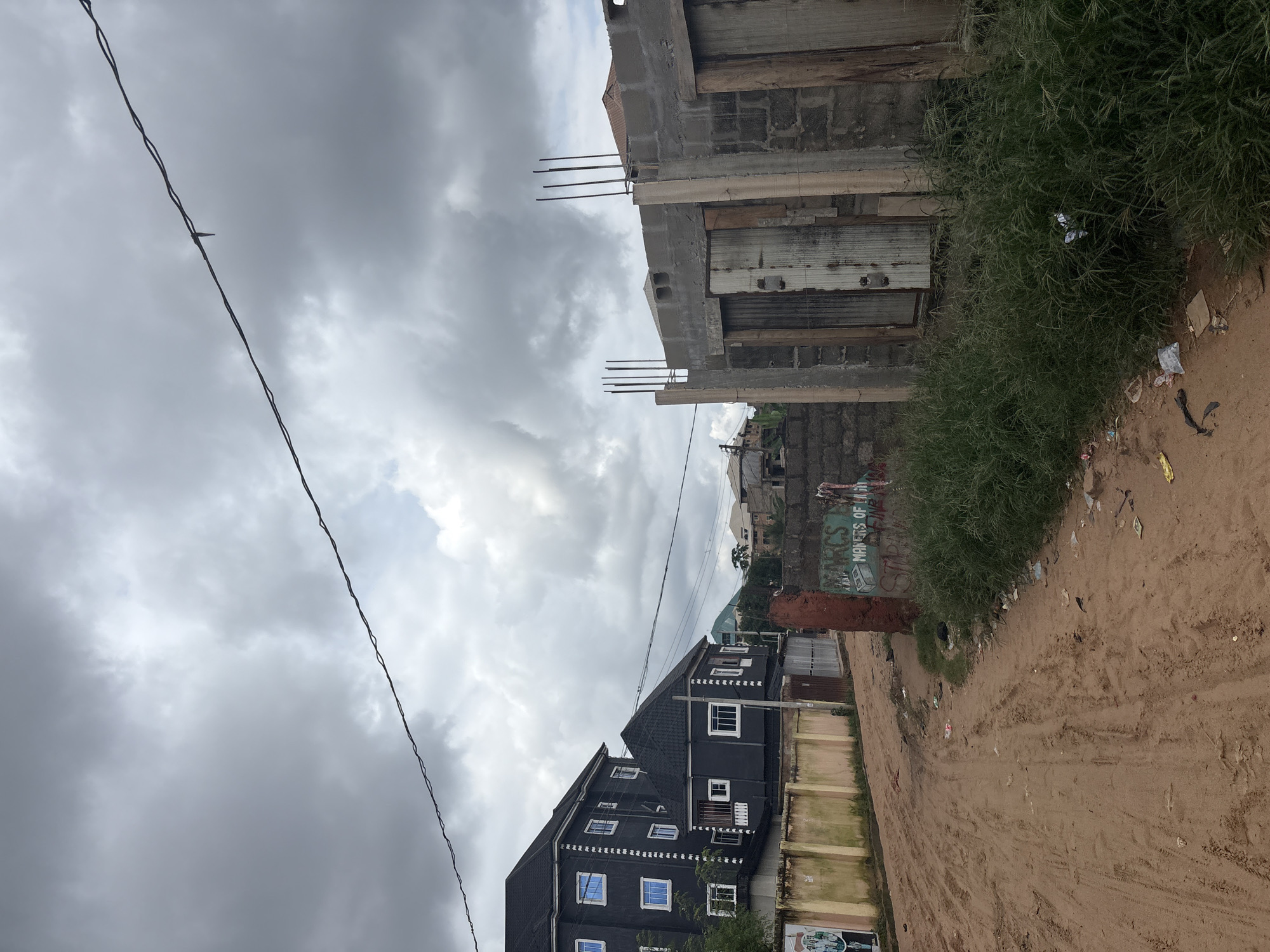 The image shows a cloudy sky above a dusty, unpaved road in Nkpor, Nigeria. The road is lined with various buildings and construction sites. On the left side of the road, a dark, multi-story building with many windows is visible, followed by a section of a light-colored wall. Further down the road, the construction of a building is in progress, characterized by exposed concrete blocks, metal reinforcement bars, and wooden planks. Some doors are visible within the unfinished structure. Green grass and weeds grow along the side of the road, with scattered debris including plastic bags and paper. In the foreground of the construction site, graffiti can be seen on a red brick pillar. The overall scene depicts an urban environment with ongoing development and a somewhat unkempt appearance.