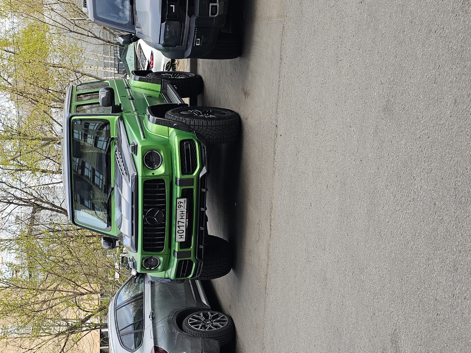 The image captures a vibrant green Mercedes-Benz G-Wagen (G-Class) parked on an asphalt surface in Khimki, Russia. The vehicle is prominently featured, facing towards the right side of the frame, showcasing its front grille, circular headlights, and distinct boxy silhouette. The hood features a dark stripe down the center.
Behind the green G-Wagen, a dark grey or black SUV, possibly a Range Rover, is partially visible. In front of the G-Wagen, the front end of a silver-grey SUV, potentially a BMW X series, is also visible. All vehicles appear to be parked on a street or in a parking area.
The scene is set during daytime with bright, clear lighting, suggesting fair weather. Bare trees with budding green leaves line the left side of the image, indicating it is springtime. Reflections of urban buildings can be seen in the G-Wagen's windows.
A notable detail is the license plate on the front of the green Mercedes-Benz, which reads "H017HH 99 RUS." The "99" is the region code for Moscow City, confirming its Russian origin as per the provided location context.