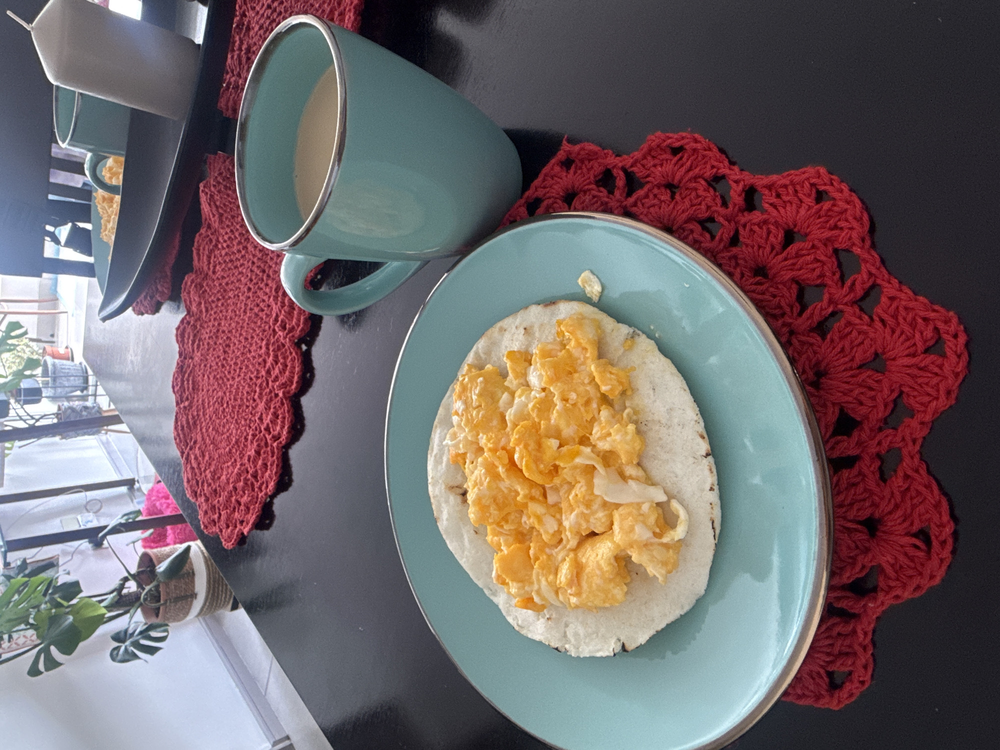 A close-up, overhead shot depicts a breakfast scene on a dark, reflective table. In the foreground, a teal plate holds a single tortilla topped with scrambled eggs. To the right of the plate, a matching teal mug is filled with a light-colored beverage, possibly coffee or milk. Both the plate and the mug rest on intricately crocheted red placemats. In the background, a black tray holds two more teal mugs and a white candle. Further back, a large potted plant with broad green leaves is visible, and beyond that, part of a doorway leading to an outdoor area. The overall impression is of a casual meal setting, likely indoors, in a residential or café environment. The city of Iquique, Chile, is not visually represented in this image.