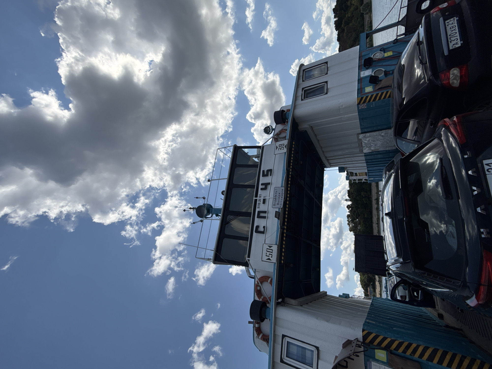 The image captures a ferry loaded with vehicles against a backdrop of a cloudy blue sky. The ferry, painted in white and teal, has the number "50" prominently displayed, along with Cyrillic text that translates to "B103B1" and "Водитель! Высади пассажиров" (Driver! Disembark passengers). The ferry's upper deck features windows and communication equipment. On the ferry deck, several cars are parked. In the foreground, a black SUV and a black sedan are visible. The sedan has a license plate that reads "A370XB 21". In the background, a lush green shoreline with trees and buildings can be seen, suggesting a town or city. The water of a river or bay is also visible. This scene is located in the city of Kozmodemyansk, Russia, on the Volga River, which is a key transportation route for ferries.