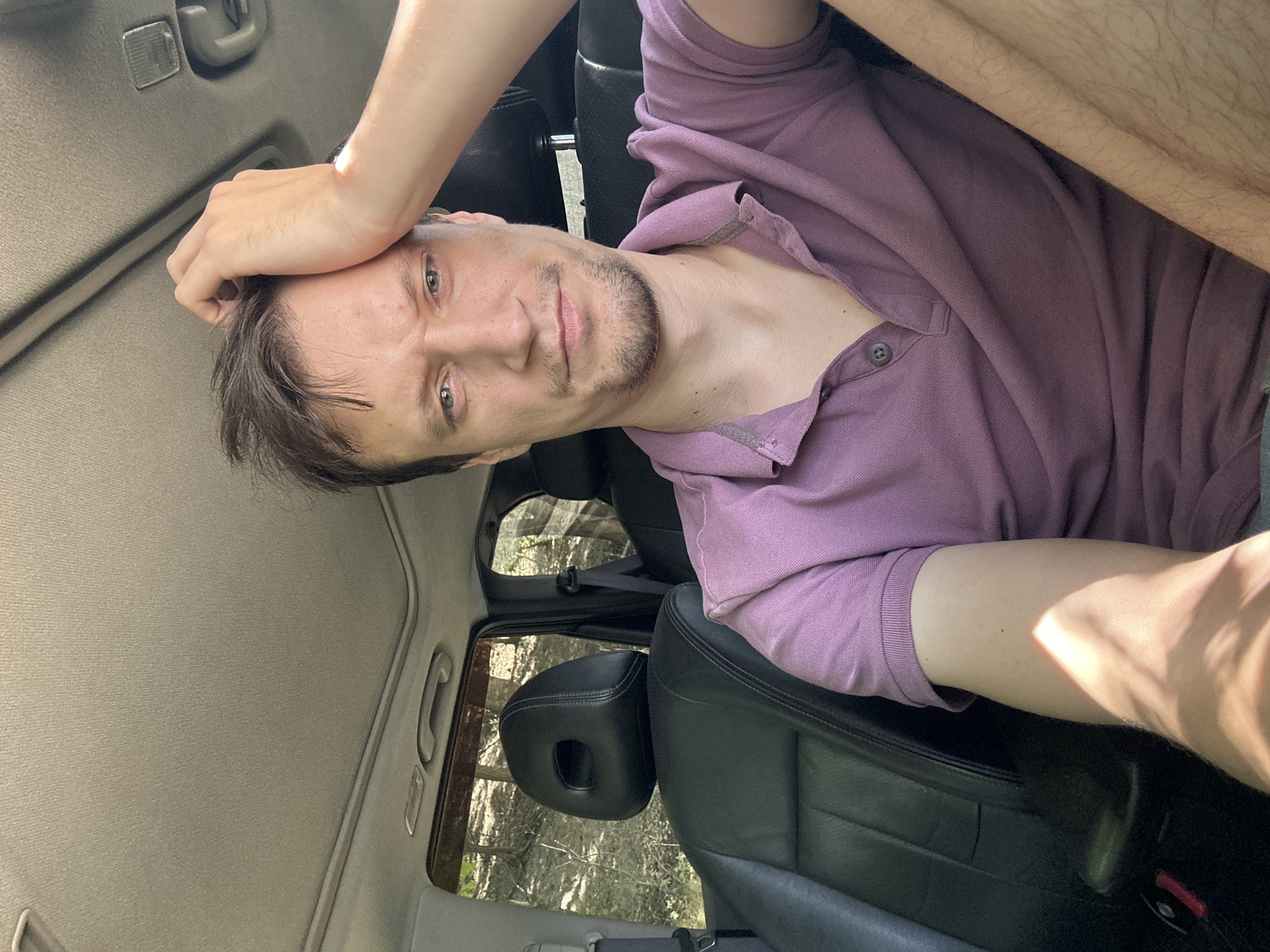 The image shows a man sitting in the driver's seat of a car. He is wearing a purple polo shirt and has his hand on his forehead. The interior of the car is visible, including the black leather seats and the sunroof. Outside the car, trees and foliage can be seen, suggesting a rural or wooded setting. The image was taken in Makiv, Ukraine.