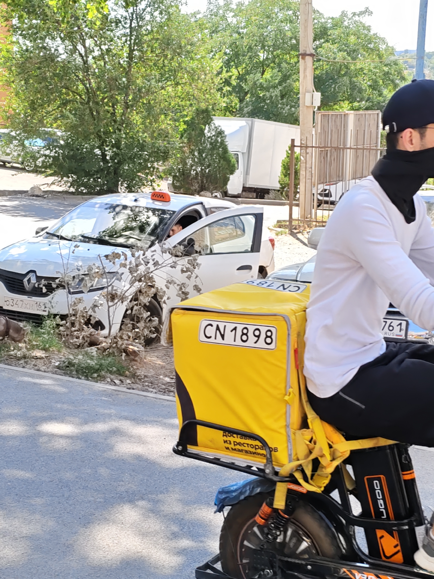 The image depicts a scene on a city street, likely in Rostov-on-Don, Russia, as indicated by a license plate on a white car. A person, wearing a black baseball cap and a black face covering, is riding an electric scooter with a large yellow delivery box on the back. The scooter has the number "CN 1898" visible on a plate attached to the delivery box. The person is wearing a white long-sleeved shirt and black pants.
Beside the scooter, a white Renault car with a taxi light on its roof is parked. The license plate of the taxi reads "O347XH 161". The car appears to be slightly off the road, partially on a grassy area. Another white vehicle, possibly a truck, is visible in the background behind a fence. The environment suggests a sunny day with trees providing shade. The road is paved, and there is a sidewalk or curb area. The overall scene captures a moment of urban delivery logistics against a backdrop of parked vehicles and greenery.