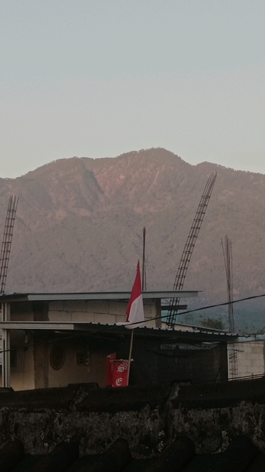 In the city of Mulyoagung, Indonesia, this image captures a landscape dominated by a large, hazy mountain range in the background. In the foreground, a dark, textured rooftop serves as a base. Above this rooftop, a two-story building structure is visible, with the upper level featuring a dark, sloped roof. A slender flagpole stands near the front of this upper level, flying a red and white Indonesian flag. To the left of the flagpole, a red flag with white lettering, possibly a banner or emblem, is also visible. Further to the left, a tall, ladder-like structure made of metal rebar rises vertically. On the right side of the frame, another metal rebar structure, partially obscured, extends upwards, and a hint of a distant, less distinct mountain or hill can be seen. The sky above is a pale, washed-out blue, suggesting either dawn or dusk. The overall impression is one of a somewhat rugged, developing urban or semi-urban environment against a natural backdrop.