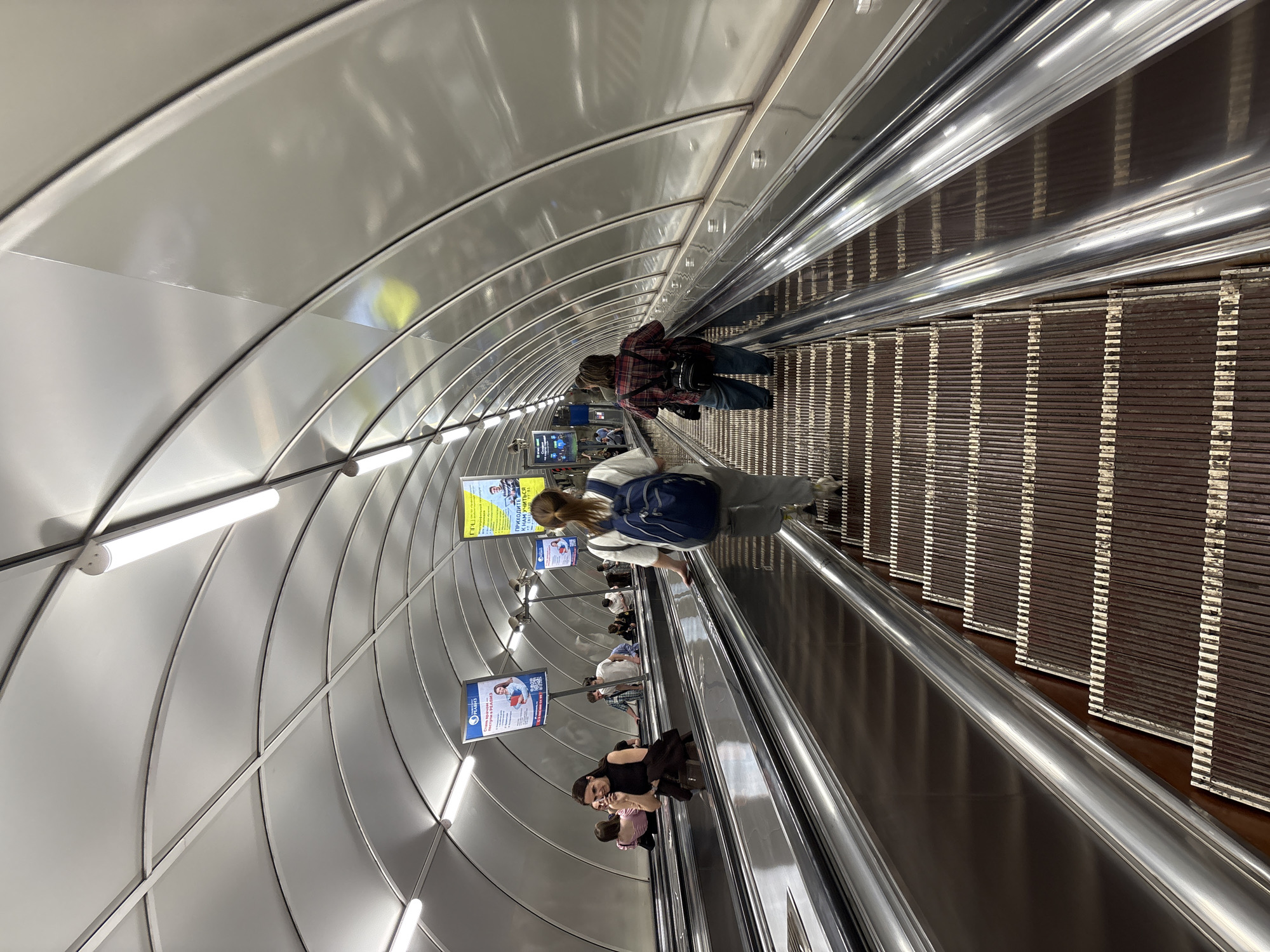 The image shows a downward-sloping escalator in an underground location. Several people are on the escalator, traveling down. The walls and ceiling are constructed of curved metal panels, illuminated by strip lights. Advertisements are displayed along the walls. The steps of the escalator are made of a dark brown material with light-colored edges. The handrails are made of a shiny, silver metal. The location is in the city of Unknown, Unknown.