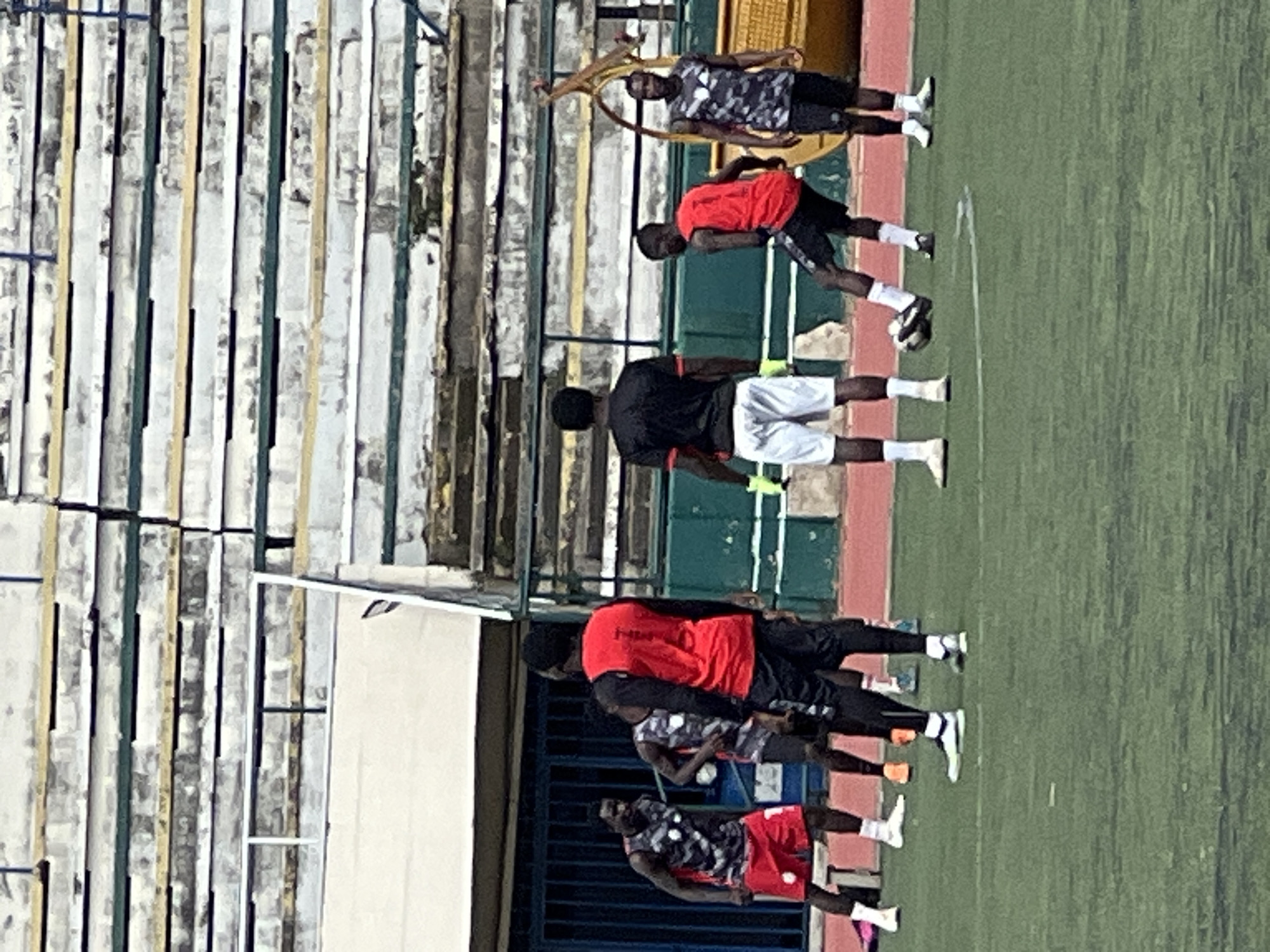The image depicts a soccer practice session at the New Stadium in Nigeria. Several male athletes, dressed in athletic wear, are engaged in drills on a green turf field. The stadium seating, a weathered structure with rows of white and beige benches, is visible in the background. Some players are in motion, either kicking a soccer ball or interacting with each other, while others are standing or observing. The lighting suggests it is daytime.