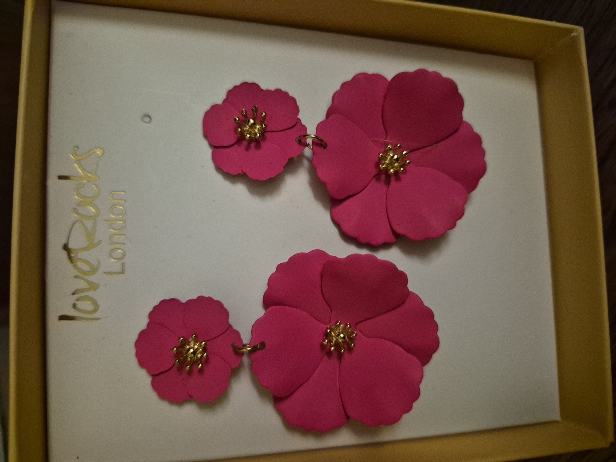 A pair of fuchsia-colored flower-shaped earrings are displayed in a white box with gold lettering that reads "loveRocks London." Each earring consists of two flower-shaped elements stacked on top of each other, with the larger flower dangling below the smaller one. Both flowers have a cluster of gold beads in the center. The box is framed by a yellow border.