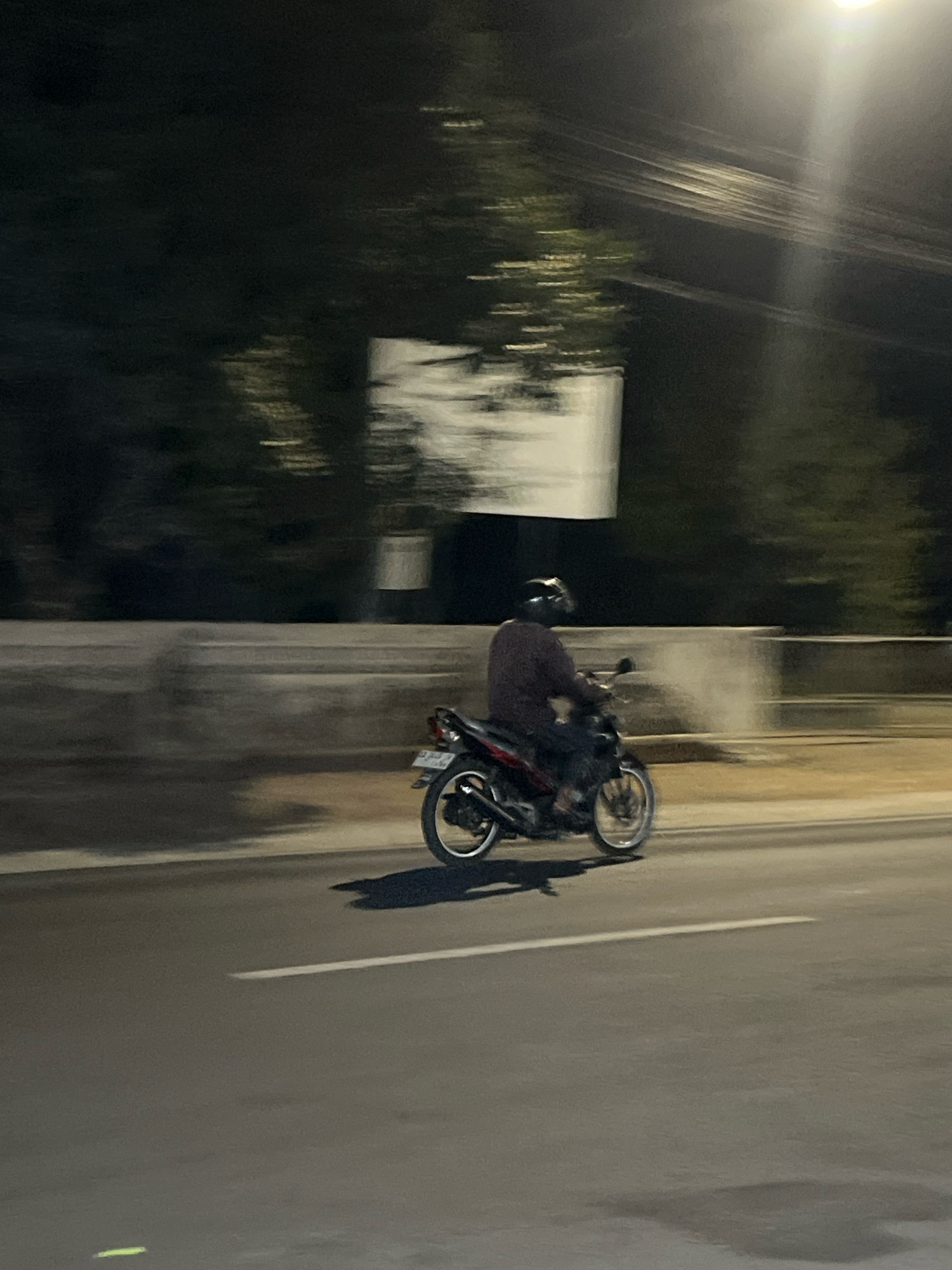 A person is riding a motorcycle at night on a road in Payaman, Indonesia. The image is blurry due to motion, suggesting the motorcycle is moving at a considerable speed. The rider is wearing a helmet and a dark-colored long-sleeved shirt. The motorcycle is dark with red accents and has a license plate that is illegible. The environment appears to be an urban or semi-urban setting with trees and a light pole visible in the background. The road has a white line marking. There is a concrete barrier along the side of the road, and a white rectangular sign is partially visible in the distance. The lighting suggests streetlights or a nearby light source illuminating the scene.