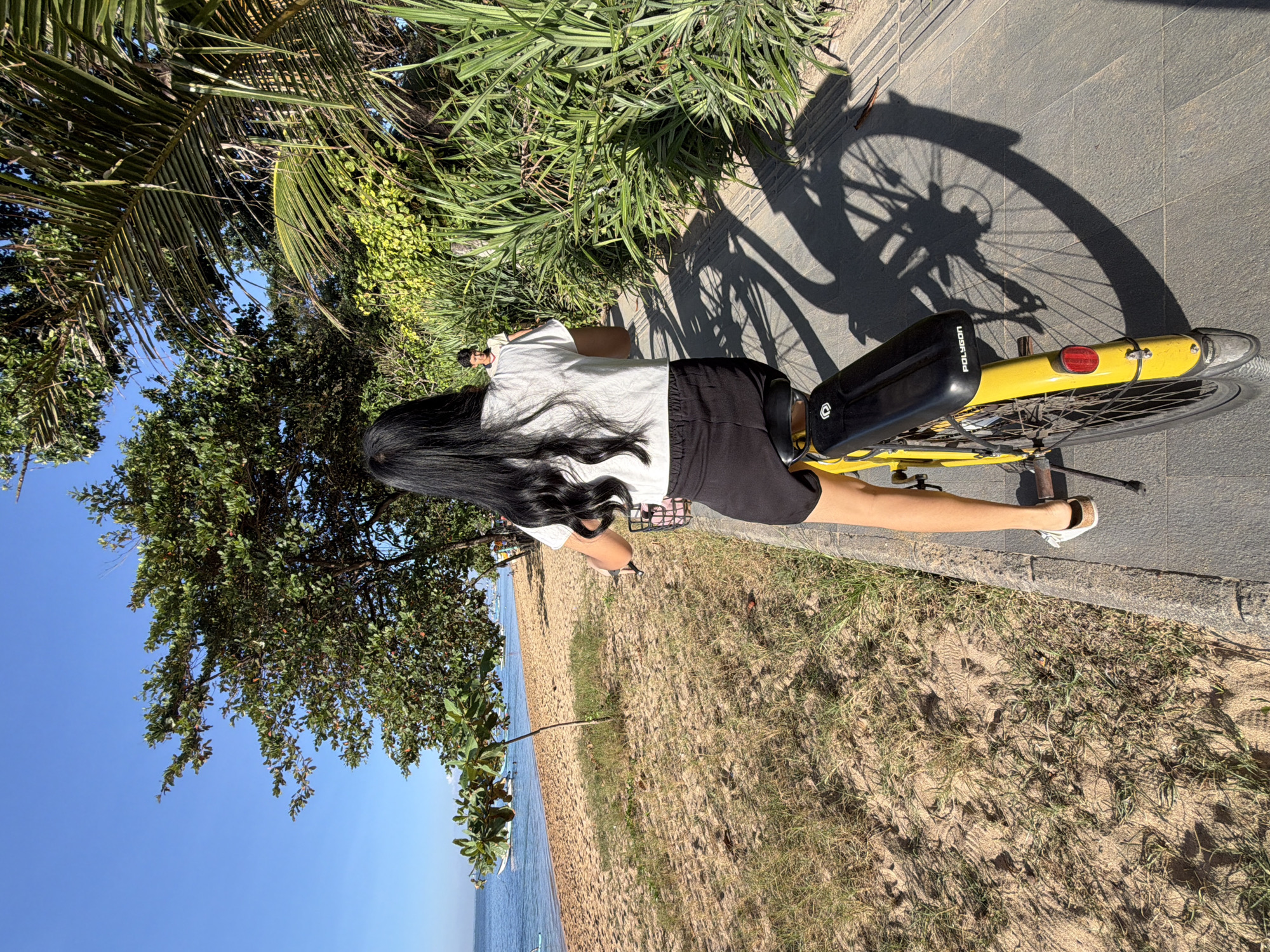 A woman with long black hair is cycling on a paved path next to a sandy beach. She is wearing a white t-shirt and black shorts, and she is riding a yellow bicycle. The beach is lined with trees and palm trees, and the ocean is visible in the background. Another person is standing in the background, partially obscured by foliage. The sun is shining, casting shadows on the path. This scene is likely located at Seminyak Beach in Denpasar, Indonesia, a popular tourist destination known for its beaches and coastal paths.