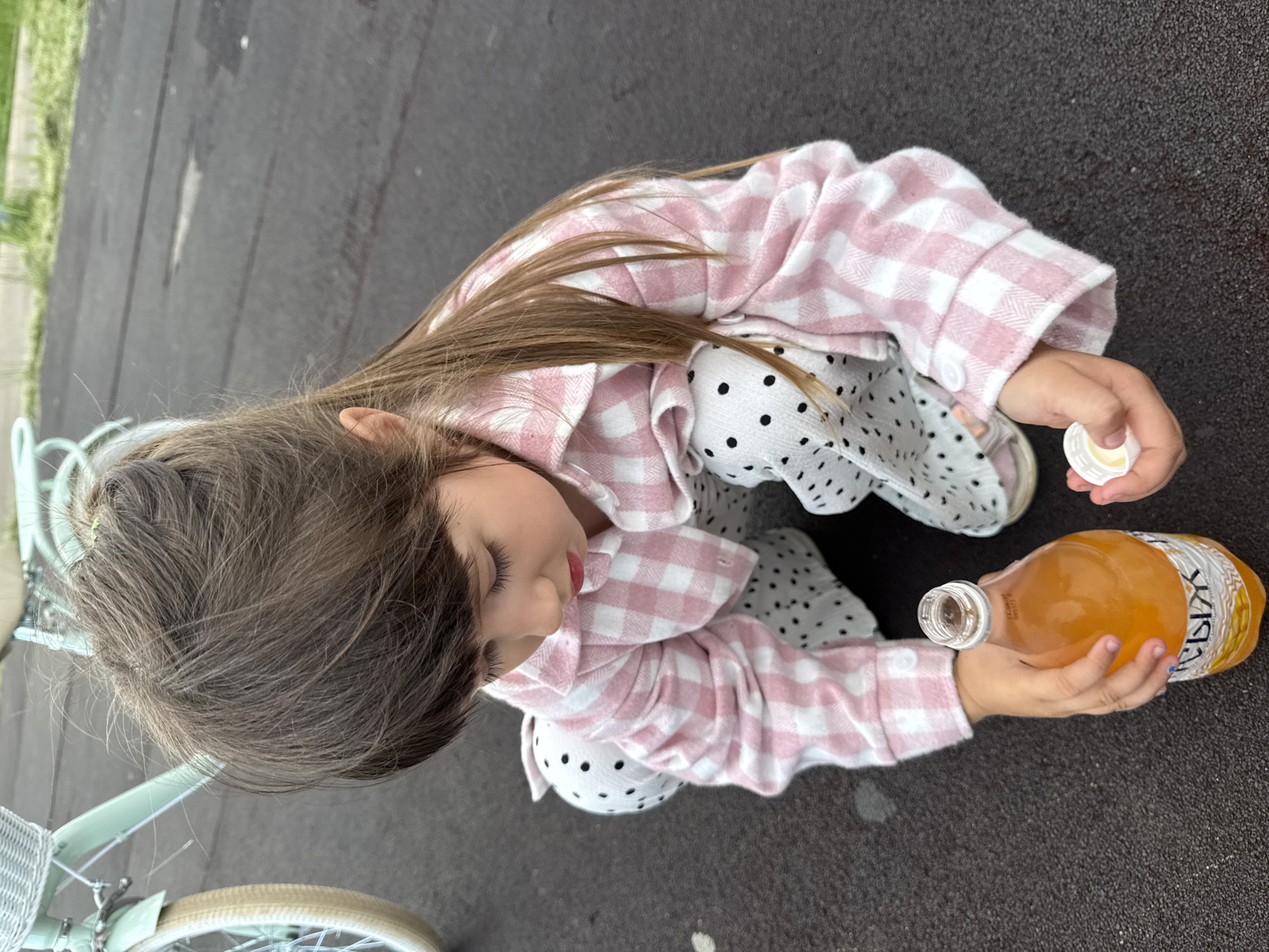 A young girl with long, brown hair is crouched down on a dark, paved surface, possibly asphalt. She is wearing a pink and white checkered shirt and white pants with black polka dots. In her right hand, she holds the white cap from a plastic bottle. Her left hand is reaching for an open, clear plastic bottle filled with an orange liquid. The bottle has a label with Cyrillic writing. In the background, to the left of the girl, a light blue bicycle with a white basket is partially visible. The environment appears to be outdoors, possibly a playground or park area. The location is identified as the city of Chaikovsky, Russia.