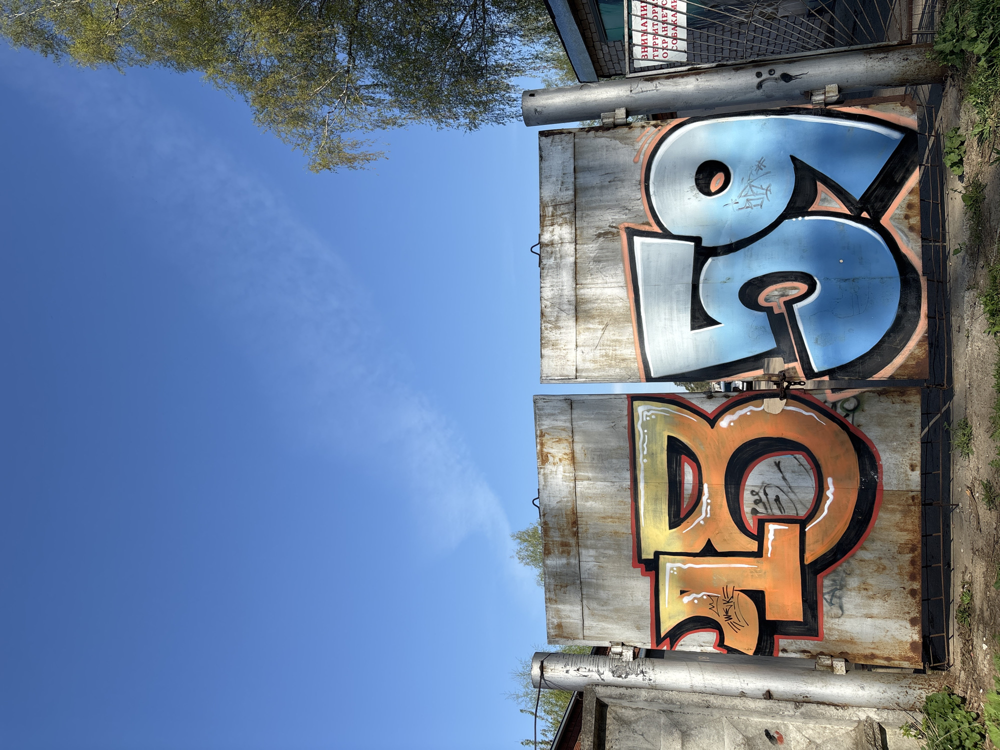 The image, taken from a low angle, shows two concrete structures with graffiti, set against a clear blue sky. A small portion of tree branches is visible at the top of the frame, suggesting the structures are outdoors. The structure on the left displays an orange and yellow graffiti design outlined in black, resembling stylized letters. The structure on the right features a blue and white graffiti design, also outlined in black, again resembling stylized letters. The surfaces of the structures are weathered and show signs of age. Based on other associated images and information, this is likely a location in the city of Chaikovsky, Russia.