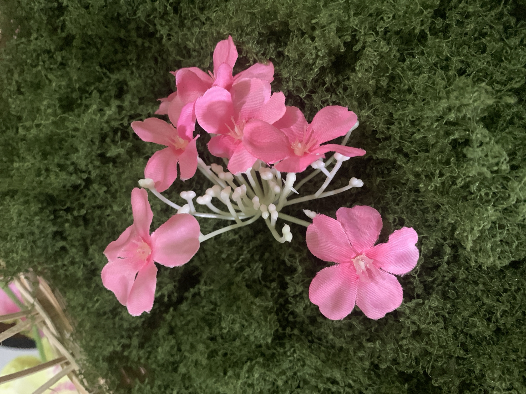 A close-up, overhead view captures a cluster of five artificial pink flowers with thin, white stems and small white buds, artfully arranged on a bed of textured, dark green artificial moss. The flowers have five petals each, and their centers are slightly darker pink. The artificial moss creates a lush, natural-looking backdrop. In the upper left corner, a portion of a light-colored, woven structure made of natural materials, possibly bamboo or straw, is visible, with a hint of pale yellow and pink in the background behind it, suggesting a decorative setting. There are no people present in the image. The scene is brightly lit, with soft shadows indicating light coming from above and slightly to the right. There is no information in the image that would allow for the determination of its location in Kuala Pembuang, Indonesia.