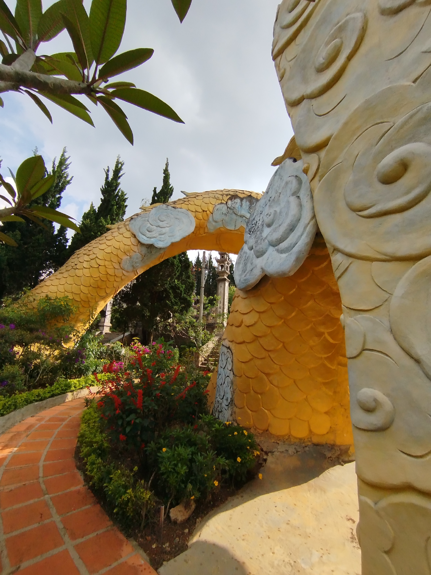 This outdoor photograph captures a decorative garden scene in Cam Ly - Đà Lạt, Vietnam, focusing on an elaborate, golden, dragon-like structure. The main subject is a large, arching form, possibly part of a dragon's body or an ornate entrance, characterized by golden, scale-like textures. Light blue-grey cloud-shaped embellishments adorn its surface. On the far right, a large, lighter-colored structure, possibly the head or another part of the dragon, exhibits intricate spiral and wave patterns.
In the foreground, lush green leaves of a tree, possibly a frangipani, extend from the upper left corner, partially obscuring the sky. A curving red-brick pathway, laid in a square pattern, winds through a vibrant garden bed in the midground. This garden features dense foliage and a variety of flowering plants, including bright red, purple, and yellow blossoms. Tall, slender coniferous trees, such as cypress or pine, are visible in the background, contrasting with the bright golden structure. Further back, hints of classical-style stone pillars can be seen, suggesting other architectural elements within the landscape.
The scene is captured during what appears to be daytime, under a partly cloudy or overcast sky, resulting in soft, diffuse lighting. There are no people or specific activities depicted. The overall atmosphere is one of serene beauty and intricate artistry within a natural setting. No visible text is present in the image.