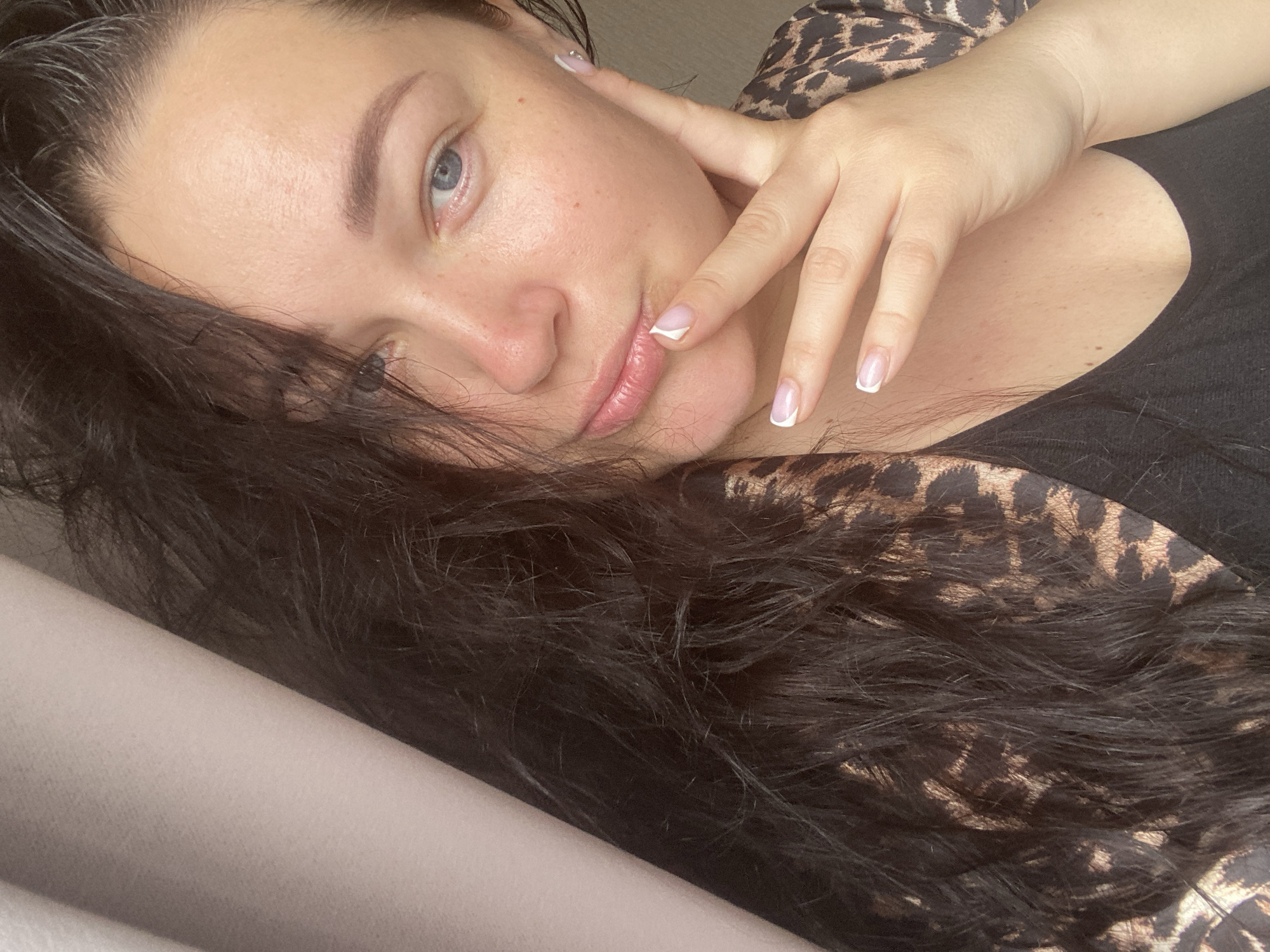 A woman with fair skin, blue eyes, and dark hair is lying down. Her right hand is touching her face near her lips. She has a manicure with white tips. She's wearing a black top with a leopard print shirt partially visible underneath. The background appears to be a light-colored surface. There's no information to ascertain the location in the city of Sochi, Russia.