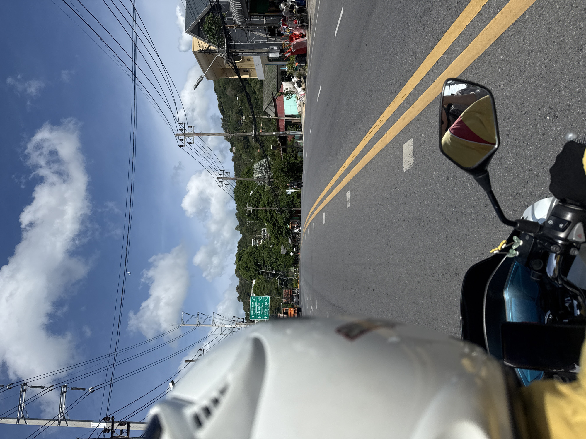 The image is a point-of-view shot from a motorcycle on a road in Karon, Thailand. A white motorcycle helmet is visible in the foreground, on the left side of the frame. The road is paved with asphalt and has double yellow lines marking the center. The sky is blue with scattered white clouds. On the right side of the road, there are buildings, some of which appear to be shops or houses, with balconies and awnings. There are utility poles and numerous electrical wires strung across the scene. In the distance, there are green, tree-covered hills. A green road sign with Thai writing and directional arrows is visible on the left side of the road, indicating directions to various locations including "Phuket Town". A truck is visible behind the sign. A motorcycle rider is reflected in the motorcycle's side mirror, wearing a yellow and red shirt. Several people are visible on the right side of the road, near the buildings. The overall scene depicts a typical sunny day on a road in a populated area of Karon, Thailand.