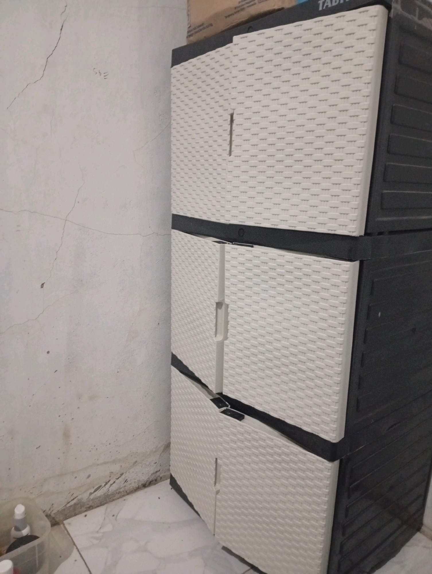 In Purwajaya, Indonesia, an indoor scene depicts a three-drawer plastic storage cabinet, primarily beige with dark grey trim, positioned against a cracked, off-white wall. The cabinet's drawers have a woven texture. To the left of the cabinet, a clear plastic container with some toiletries is visible on a tiled floor. The wall displays evidence of water damage and staining near the floor. A small, handwritten marking, possibly "395" or "Sg5", is present on the wall above the water damage.