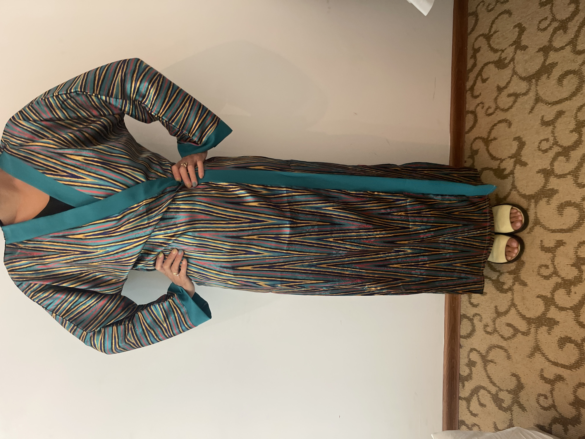 A person stands against a white wall in what appears to be a room. They are wearing a long, flowing robe with a colorful, vertical stripe pattern in shades of blue, yellow, pink, and dark brown. The robe has a wide, teal-colored lapel and trim. The person has their hands on their hips, wearing gold sandals. The floor is carpeted with a tan and brown swirling pattern.