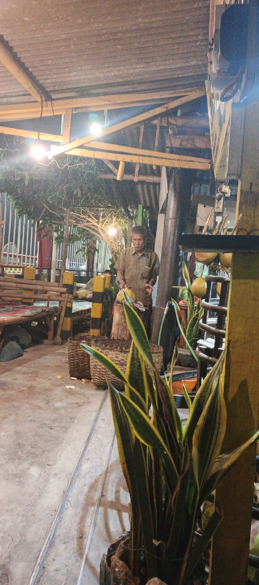 An overhead shot shows a nighttime scene of an outdoor food stall in Kasiyan, Indonesia. A man in a green shirt is standing and preparing food. In the foreground, a potted snake plant with green and yellow leaves is visible. In the middle ground, two woven baskets sit on the ground. In the background, a bamboo fence and a white metal fence enclose the area. Several light bulbs illuminate the scene, creating shadows and highlights on the concrete ground. A person can be seen resting on a bench in the distance.