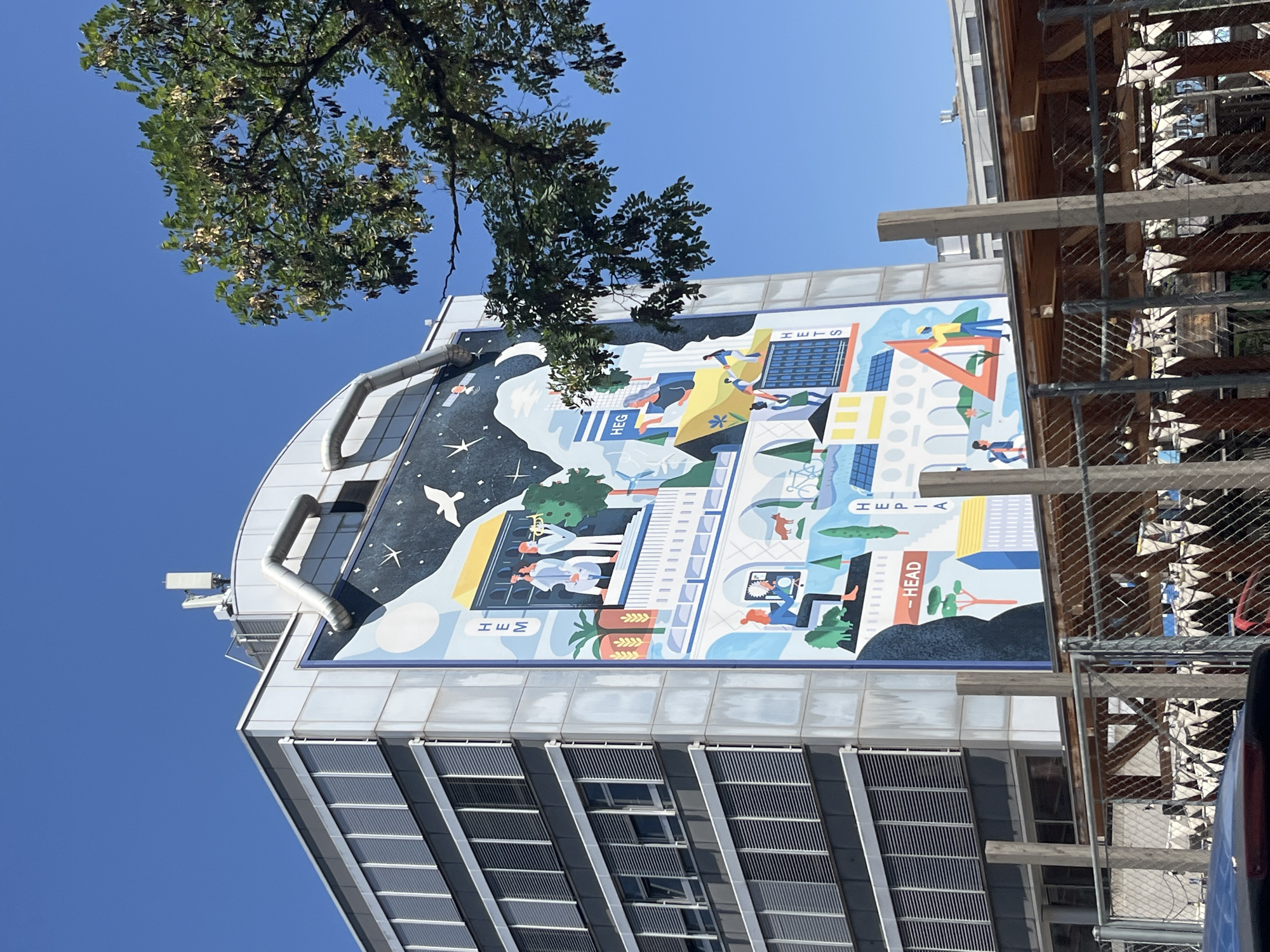 The image shows a mural on the side of a modern building with a curved facade. The mural depicts a stylized representation of urban life and technology. In the upper left quadrant of the mural, a night sky with stars and a moon is illustrated, with a white bird in flight. The rest of the mural is a vibrant cityscape with colorful buildings, trees, and abstract shapes. Various scenes of people engaged in different activities are depicted, such as people working on computers, riding bicycles, and interacting with each other. There are also symbols of technology like solar panels and a laptop. Text within the mural includes "HEG," "HETS," and "-HEAD." The building itself has a sleek, contemporary design with horizontal slatted windows. In the upper part of the image, a tree with green leaves partially obscures the sky, which is a clear blue, suggesting it is a sunny day. The building appears to be located in an urban environment, possibly a university campus given the "HEG" and "HETS" text, which could refer to an institution. Part of a wooden structure and a chain-link fence are visible to the right of the building, suggesting ongoing construction or maintenance.