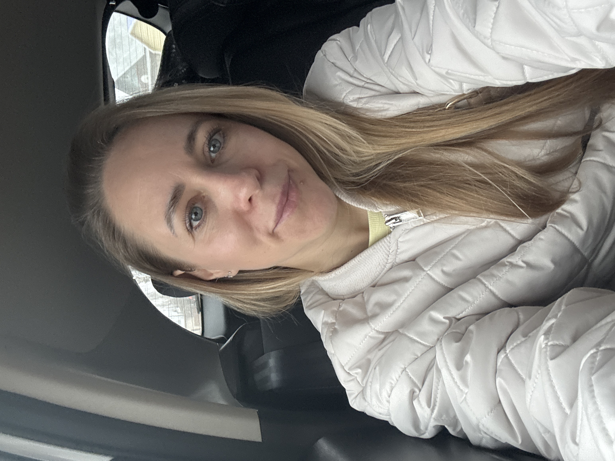 A woman with blonde hair and blue eyes is sitting in a car. She is wearing a light-colored quilted jacket and a yellow top. The car interior is dark. The car window shows a blurred outdoor scene that may be in an urban area. Based on the information available, I cannot confirm this image was taken in the city of Chaikovsky, Russia.