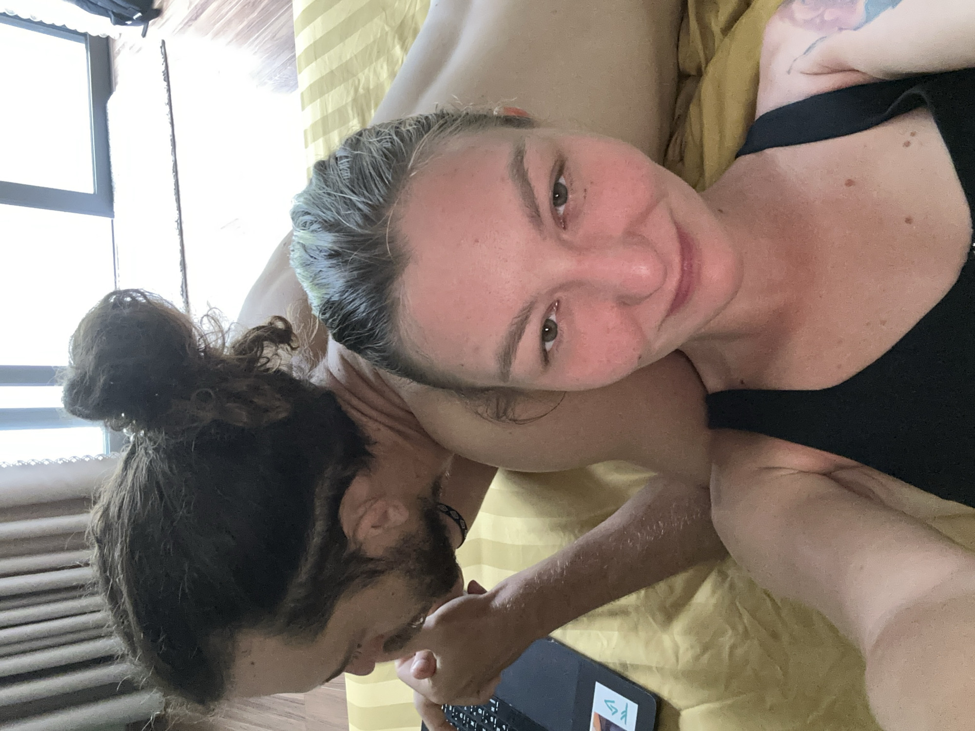 A woman and a man are lying together on a bed with a striped yellow sheet. The woman is in the foreground, looking at the camera with a slight smile. She has short, light hair and is wearing a black tank top. Her right arm, featuring a tattoo, is visible. The man is behind her, with his head near her shoulder. He has long, dark hair pulled back in a bun and a beard. He is holding a tablet with his right hand. The bed is near a window with vertical blinds. The location is described as Hà Giang 1 Ward, Vietnam.