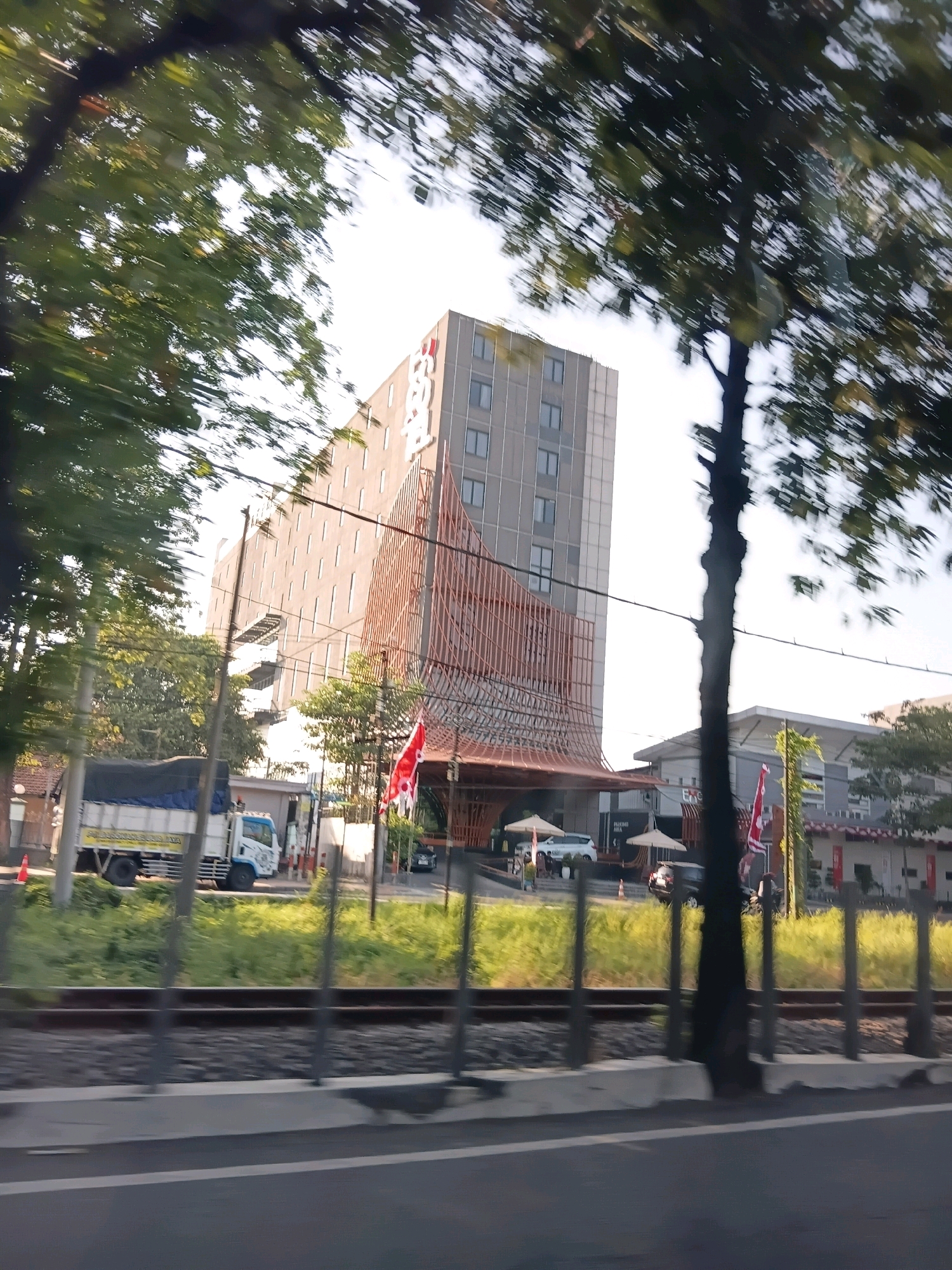 This is a photograph taken from a moving vehicle, likely on a road adjacent to a railway line, in the city of Surabaya, Indonesia. The main subject is a modern, multi-story building with a distinctive architectural design featuring a facade of vertical wooden slats. The building appears to be a hotel or commercial establishment, with the name "Four Points" visible in large lettering near the top. In front of the building, there is an entrance area with white umbrellas and several parked cars.
To the left of the building, a white truck with a blue tarp covering its cargo is parked on the side of the road. The truck has some text on its side, but it is illegible. Further to the left, there are trees with green foliage, some of which are partially obscuring the view of the building and the sky.
The foreground of the image shows a road surface and a concrete barrier. Behind this, a set of railway tracks is visible, with ballast and sleepers. The overall scene suggests an urban environment with a mix of modern architecture, transportation infrastructure, and natural elements. The lighting indicates it is daytime, with bright sunlight casting shadows. The motion blur suggests the photograph was taken while the vehicle was in motion.