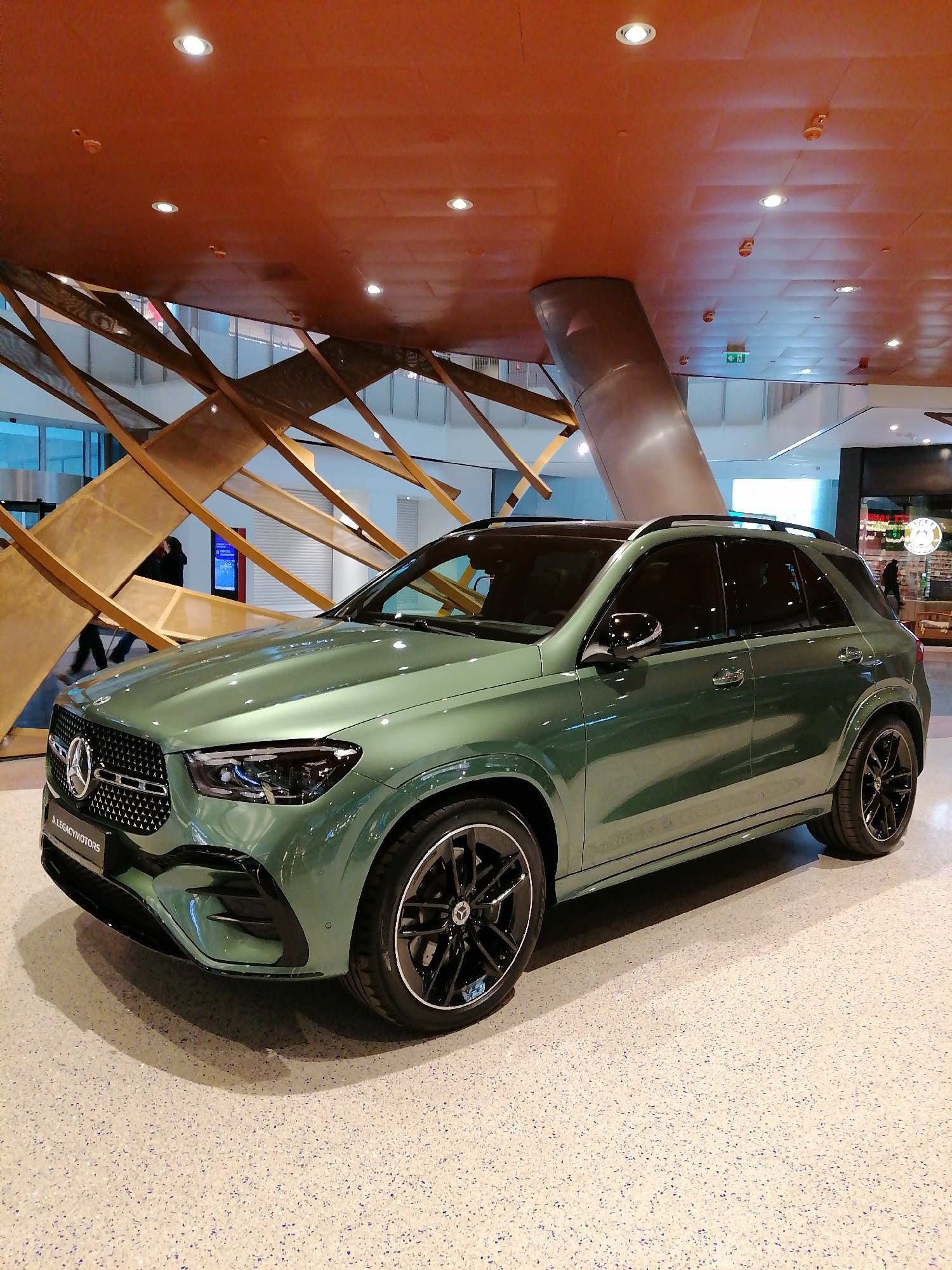 The image features a green Mercedes-Benz GLE SUV displayed indoors, likely in a dealership or showroom. The car is positioned on a light-colored speckled floor. In the background, there's a complex wooden structure, possibly part of the building's architectural design. A cylindrical pillar and a large orange-toned ceiling with recessed lighting are also visible in the background. The location is in the city of Moscow, Russia based on the license plate reading of the car, suggesting that the vehicle is local to Russia.